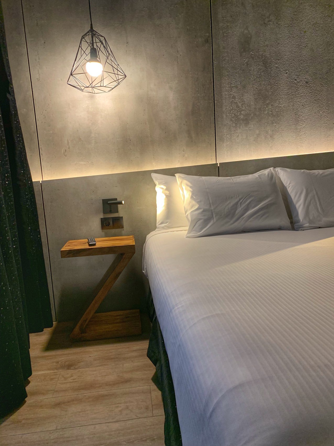 A bed against a concrete wall under a wire pendant lamp, with a green curtain beside it
