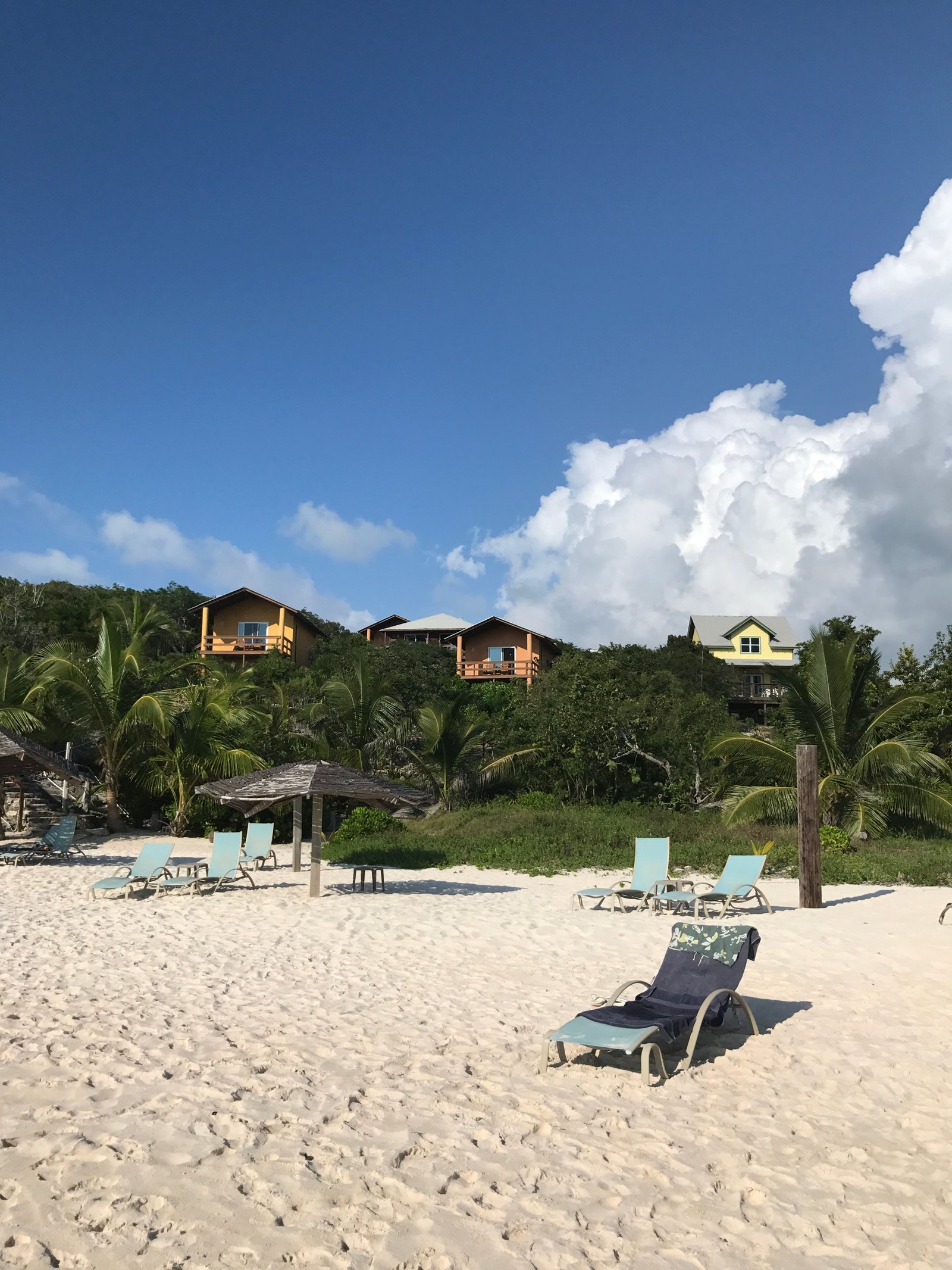 Loungers and a palm thatch shade on white sand, with ochre and yellow cottages on the wooded hill above