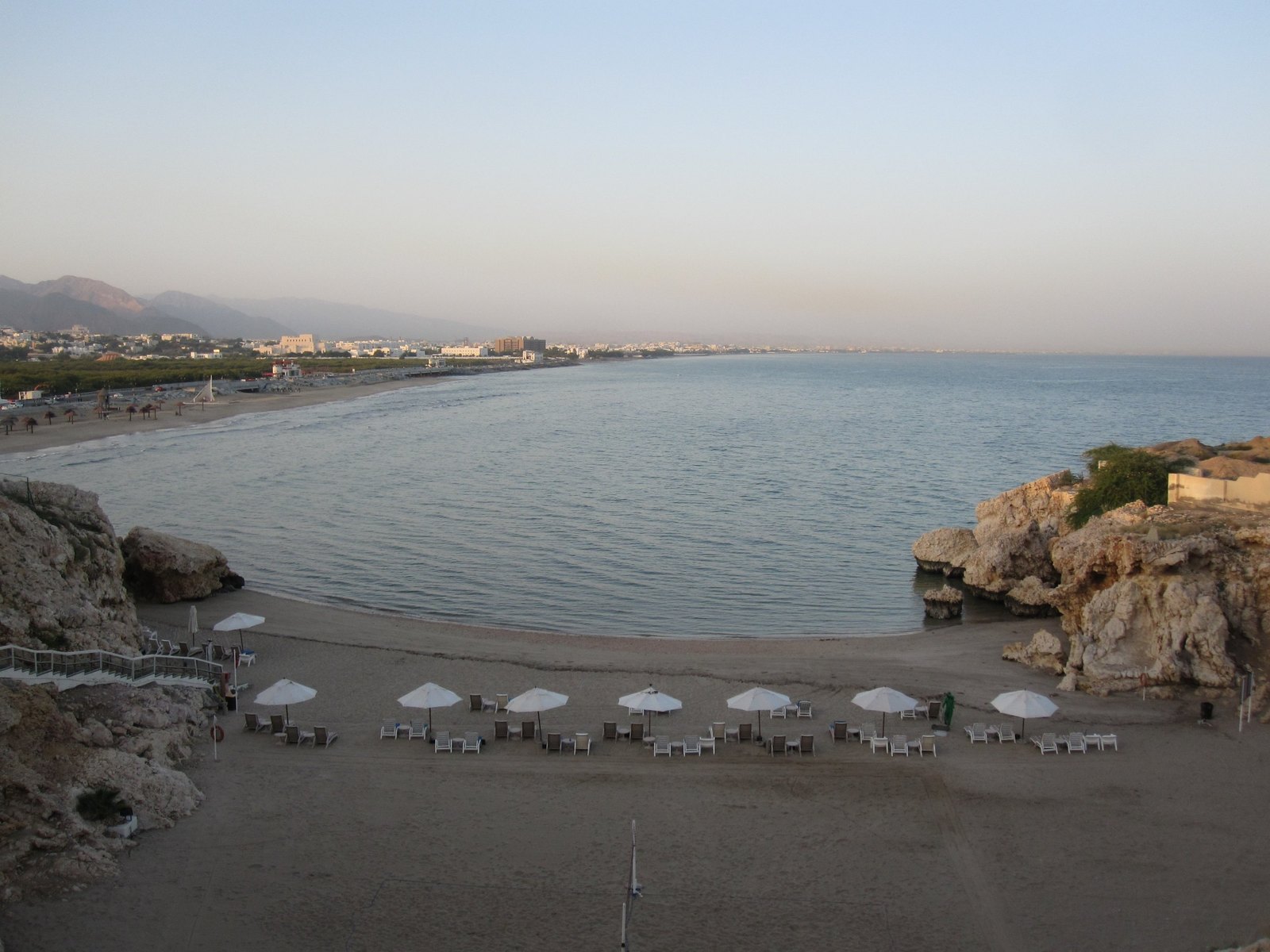 A sand cove between two rock headlands seen from above, parasols in a line and the Hajar mountains behind the town