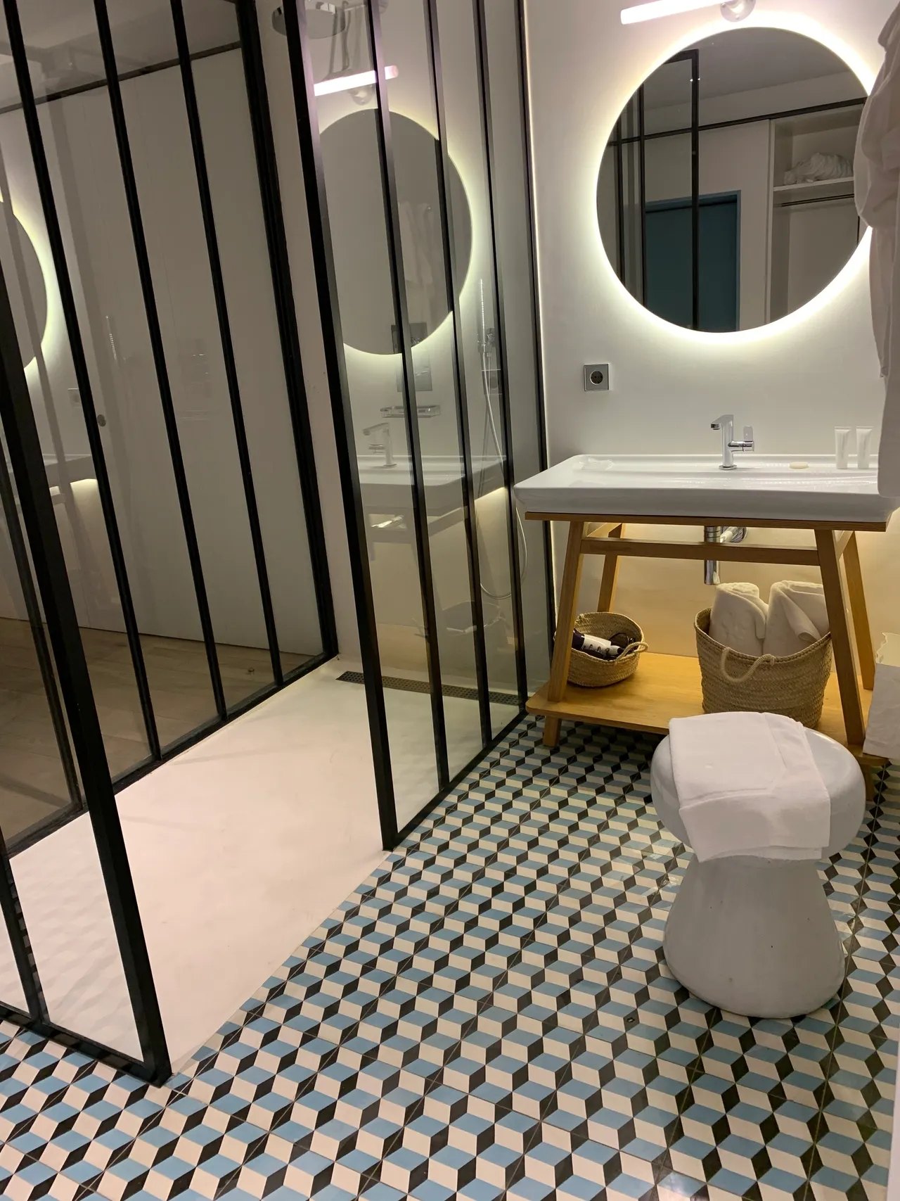 A bathroom with a black framed glass shower screen, a round backlit mirror above a white basin on a timber trestle, baskets of towels beneath it, and a floor of blue, black and white tiles laid as tumbling cubes