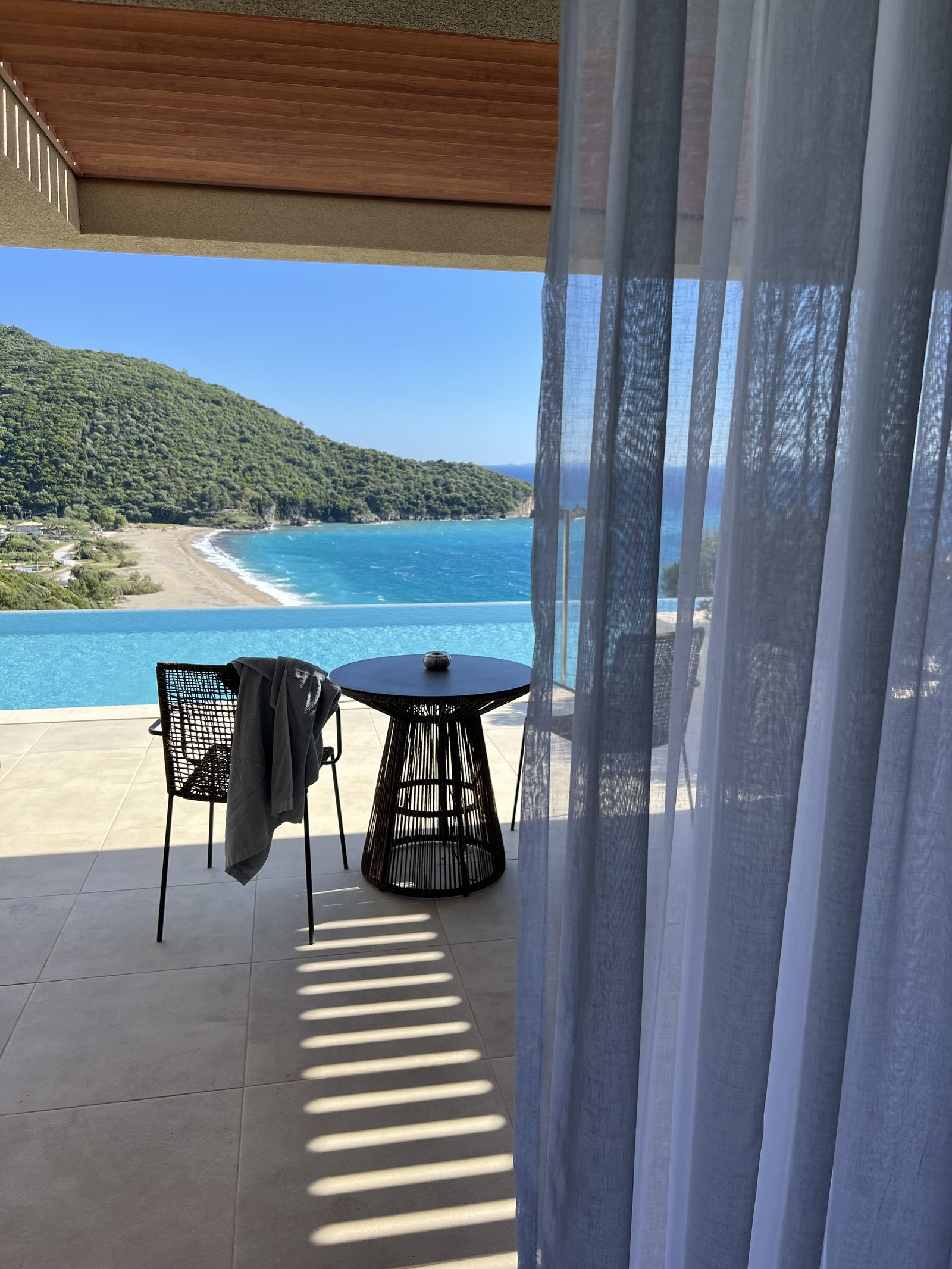 A gauze curtain drawn back at the edge of a terrace with a chair and a low table, looking over a pool and across a bay to a crescent of beach under a green headland