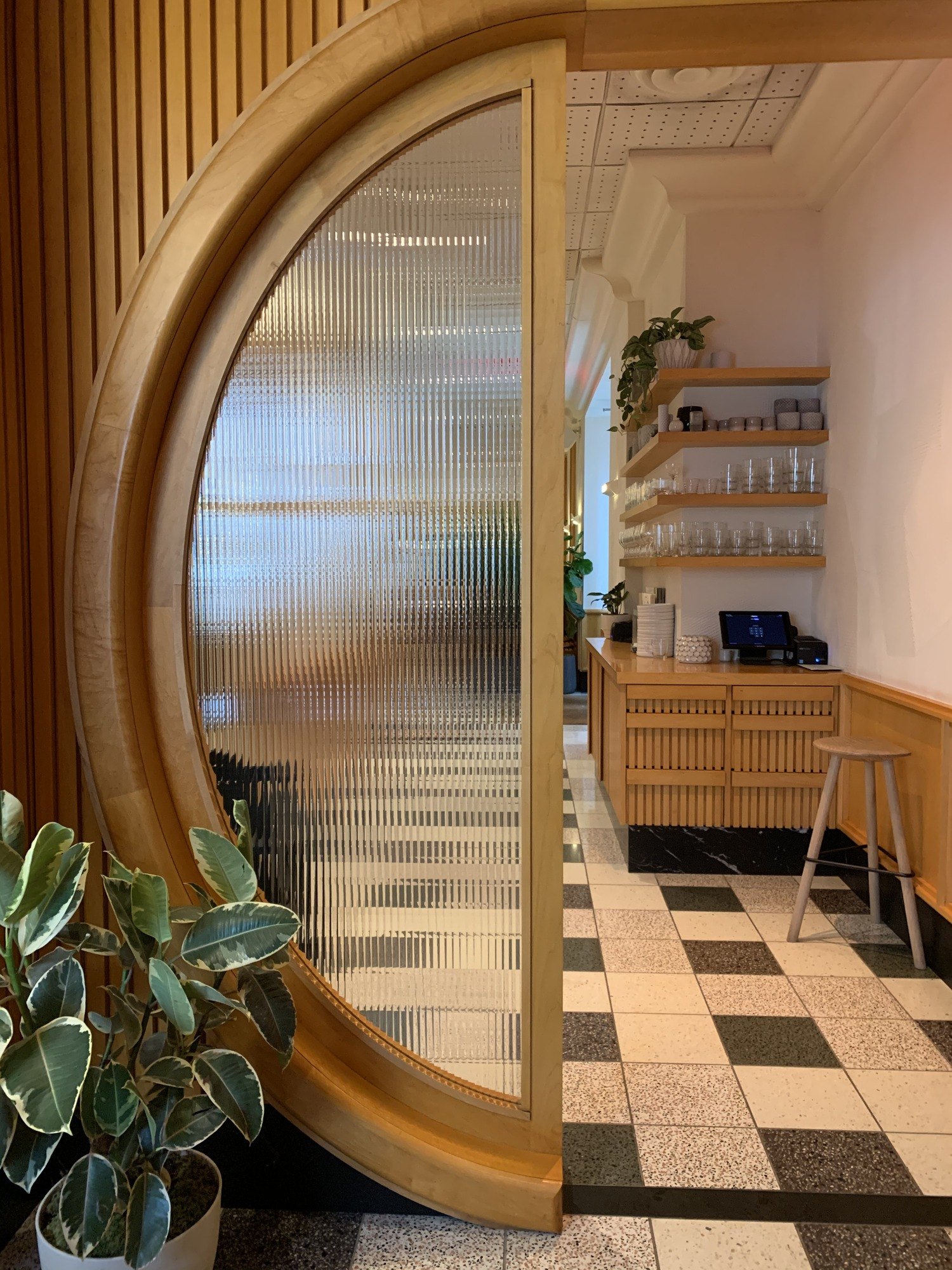 A door of fluted glass in a curved oak frame standing open onto a room of shelved glassware, with a rubber plant in the foreground and terrazzo underfoot