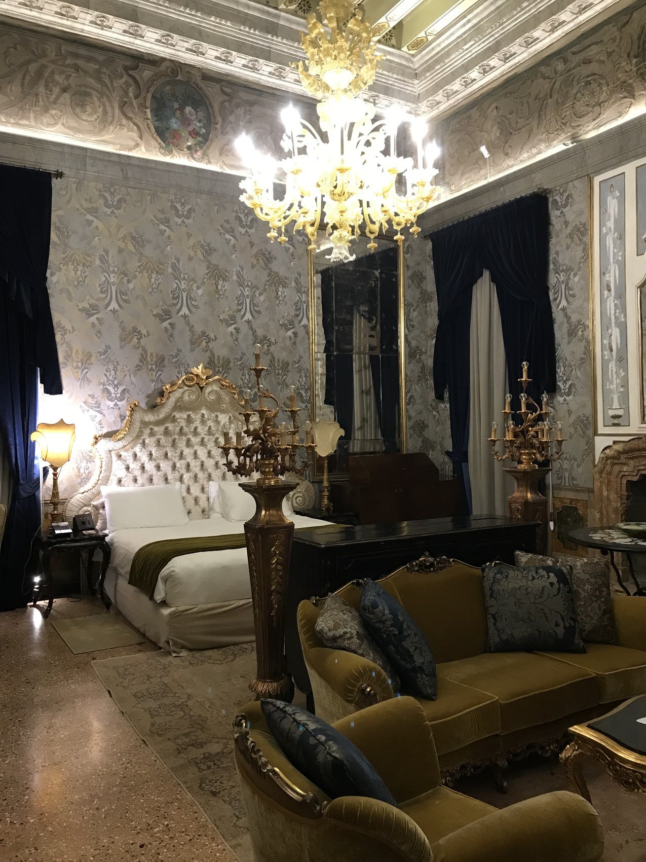 A room in silver damask with a Murano chandelier, a gilt bed and a green velvet sofa