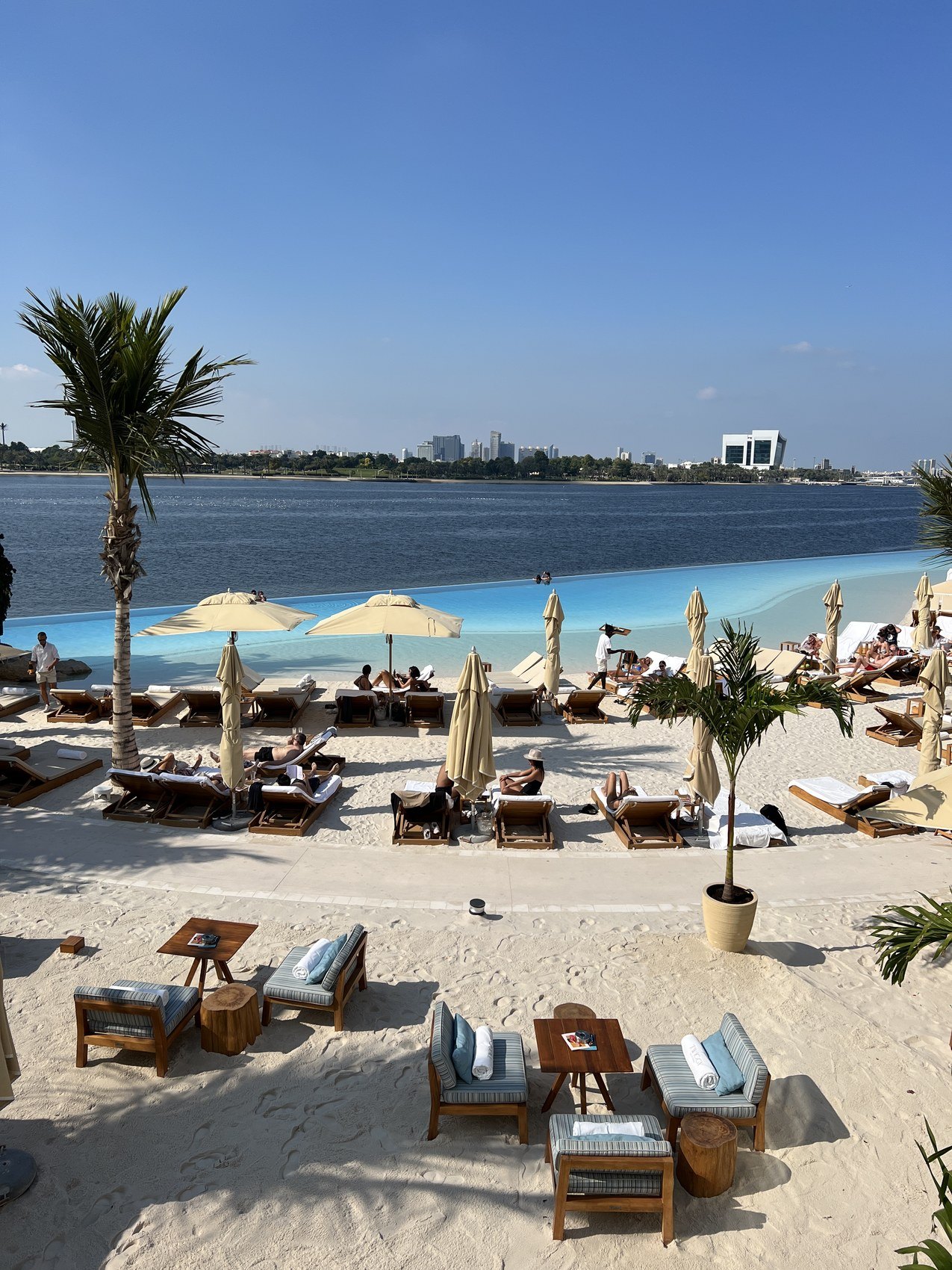 Loungers and parasols on white sand along a bay, towers on the far shore