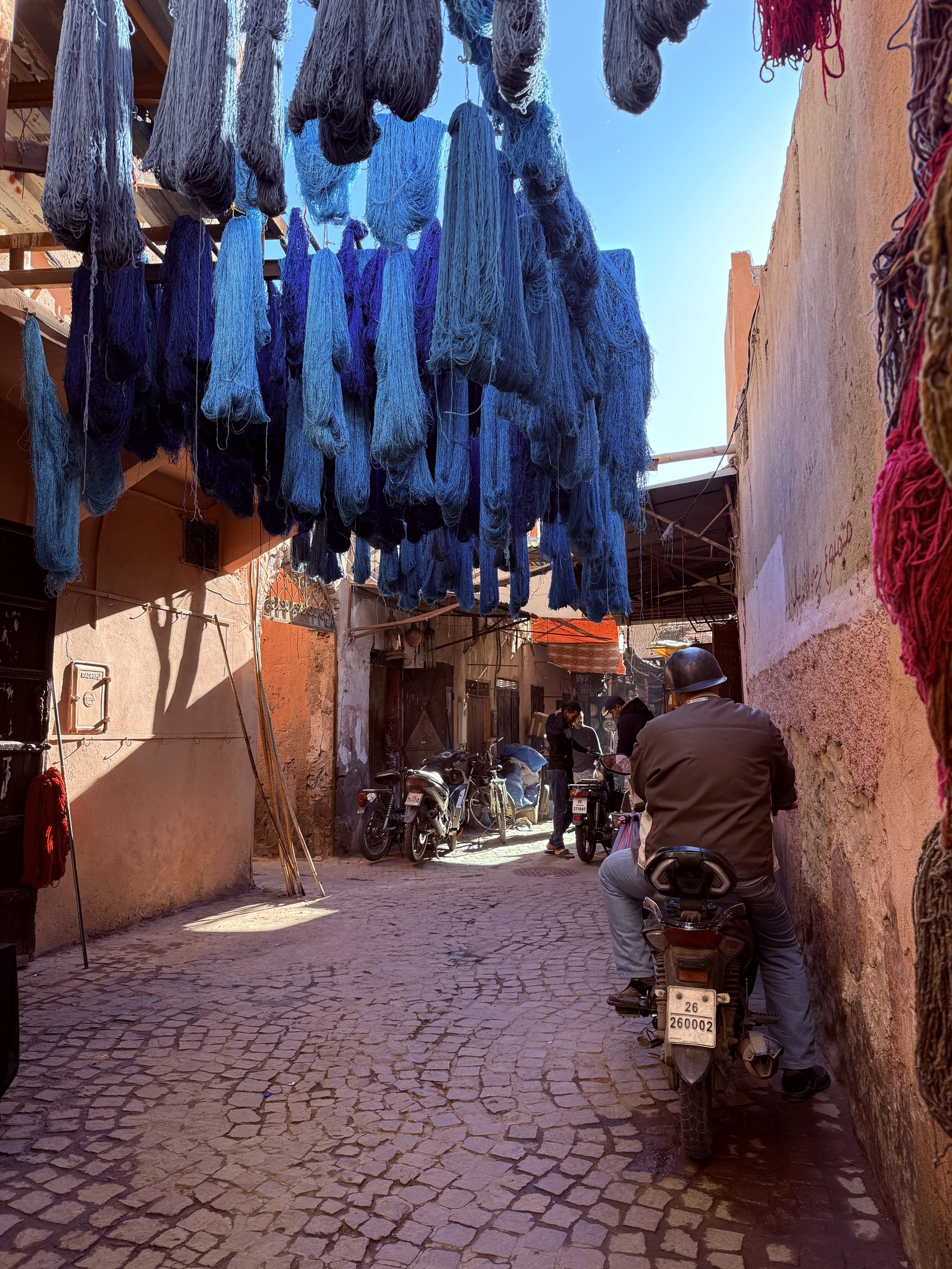 Skeins of wool dyed deep blue hanging to dry on poles above a narrow lane of the medina, with pink rendered walls, red skeins at the right and a man riding a motorbike away down the cobbles