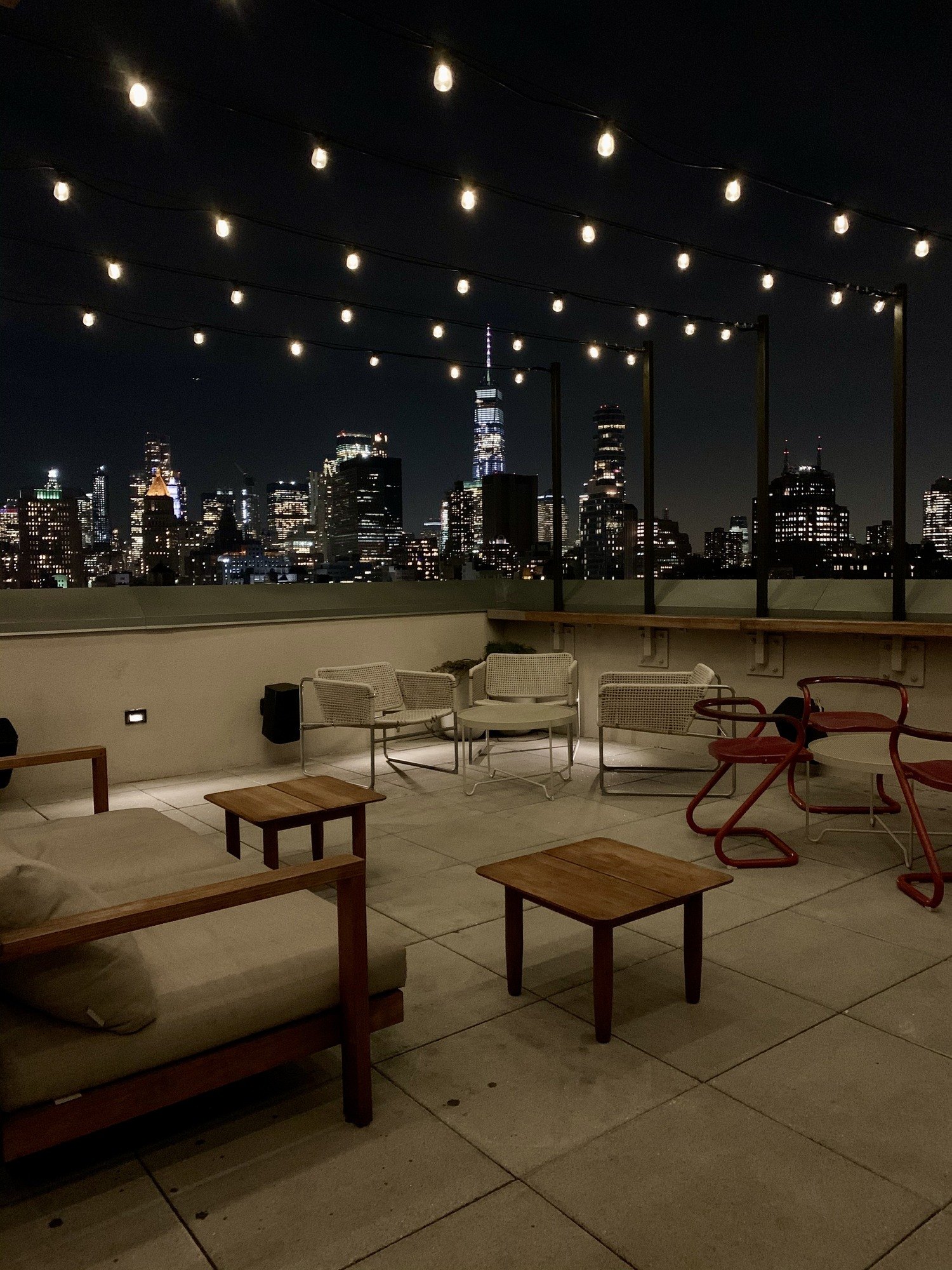 A roof terrace at night under strings of bulbs, with low timber seating and red metal chairs and the lit towers of Manhattan behind