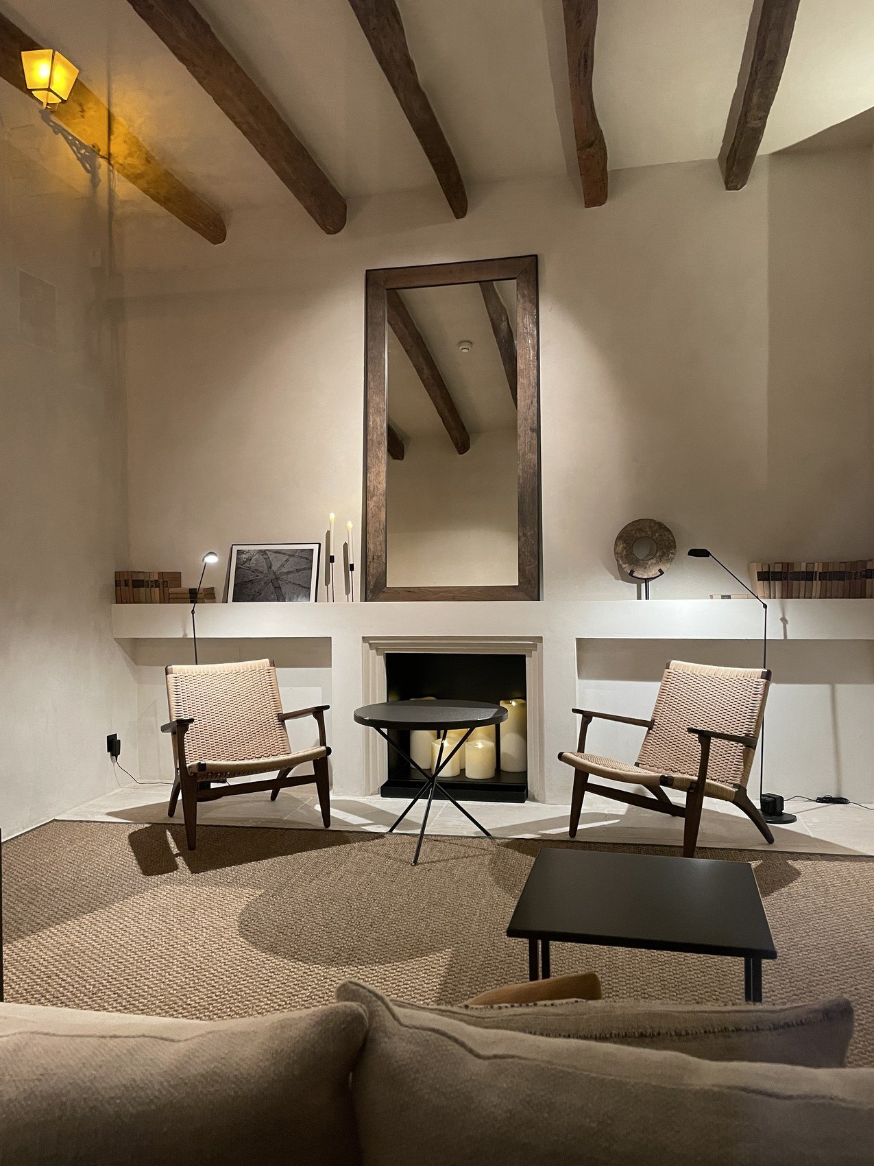 Two low chairs facing a fireplace under a white beamed ceiling, a mirror above the mantel