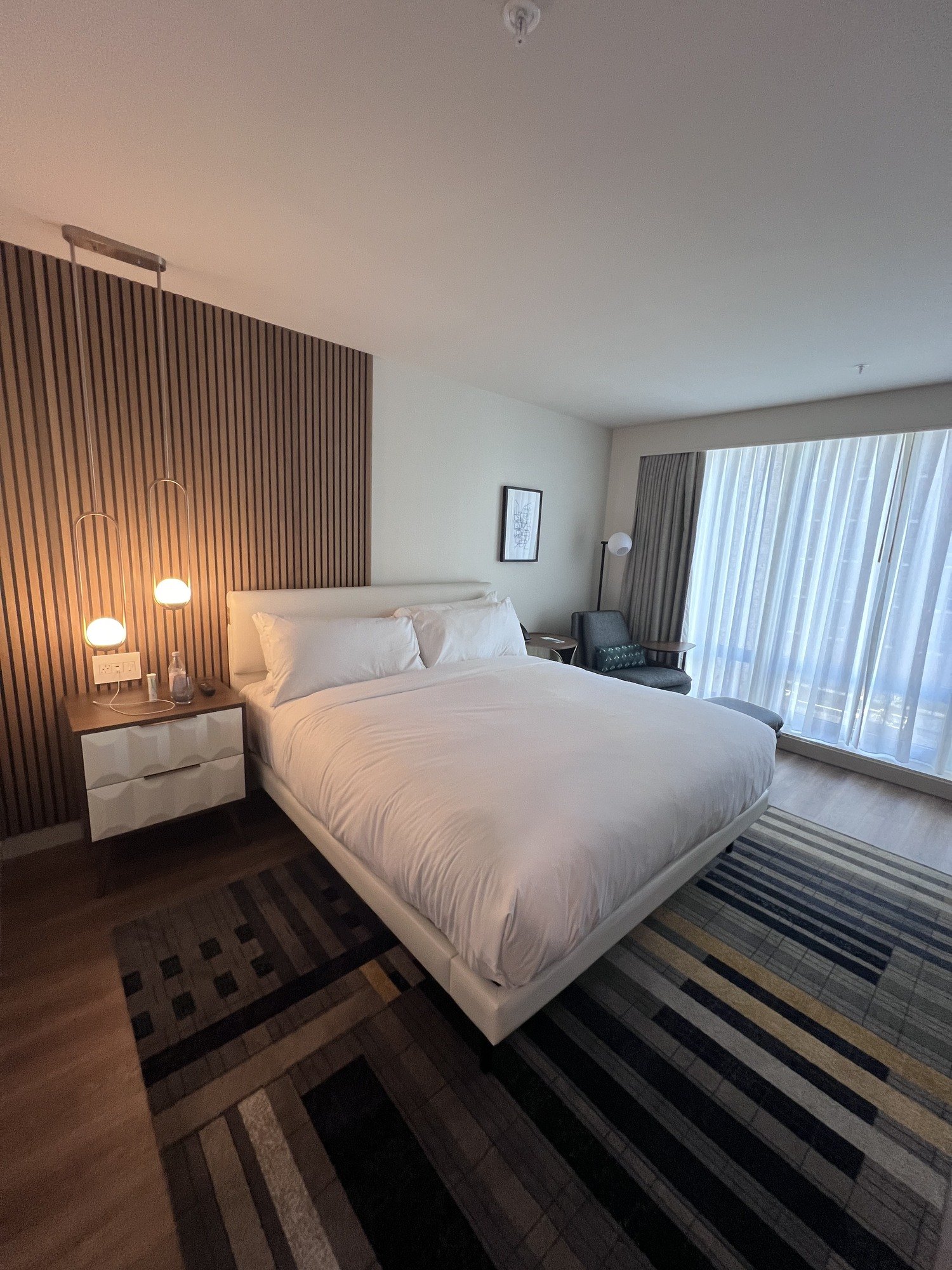 A bed with white linen against a full wall of fluted pale timber, with reading lamps either side and a striped carpet running under it to a curtained window