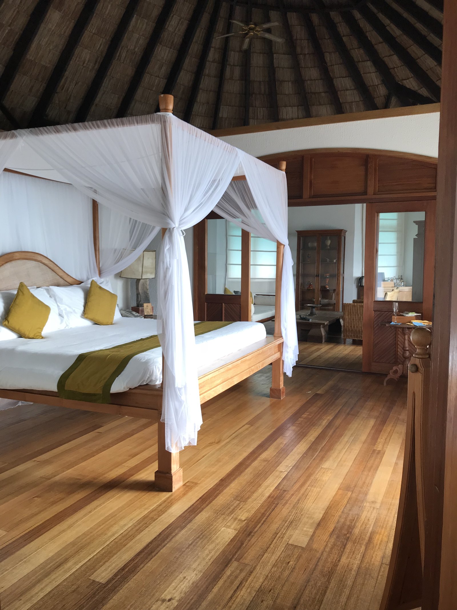 A four poster bed draped in white mosquito netting in a timber villa under a thatched roof