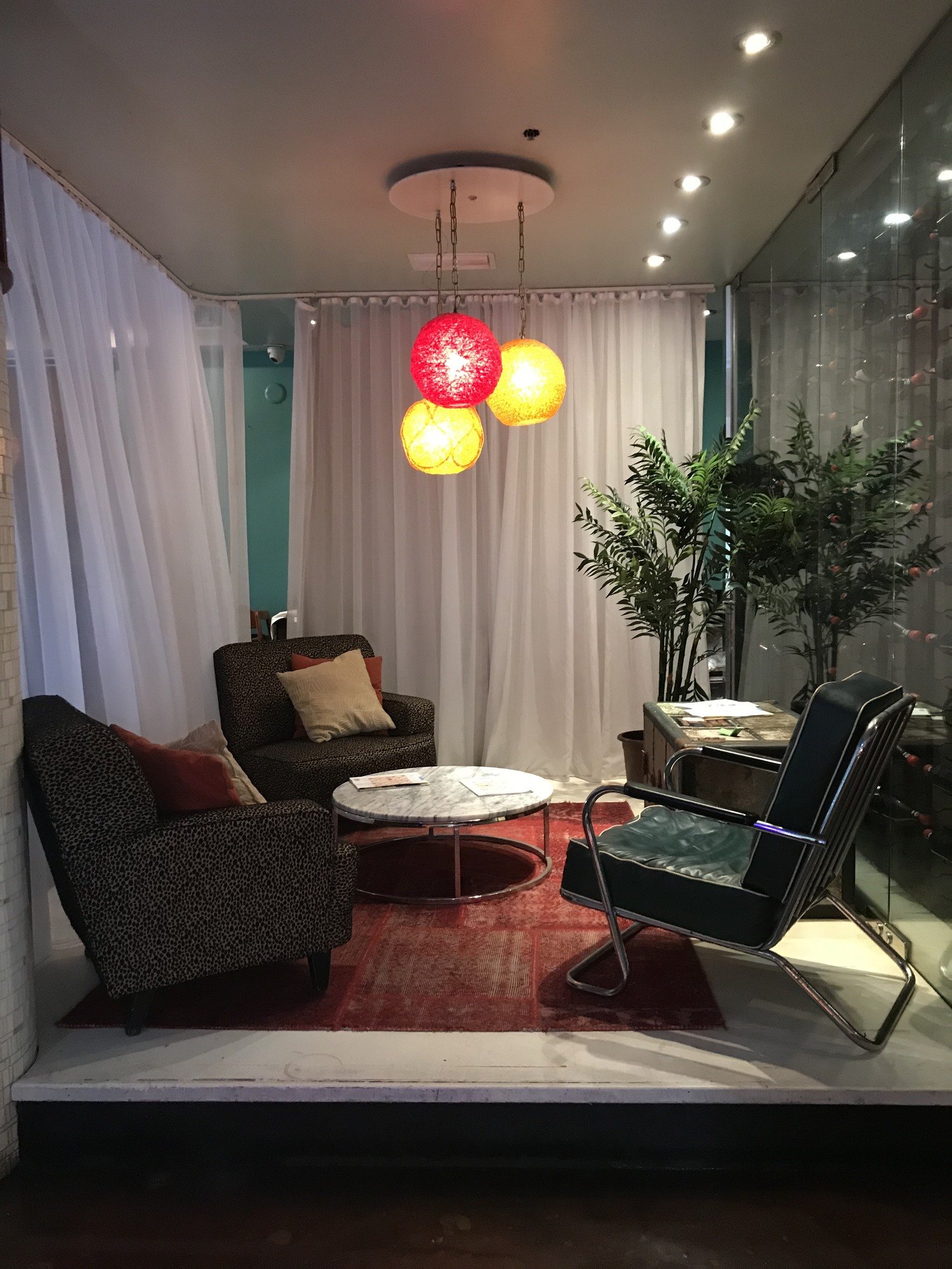 A sitting area behind a drawn white curtain with a cluster of red and yellow glass globe pendants above two lounge chairs and a round table, with plants in the corner