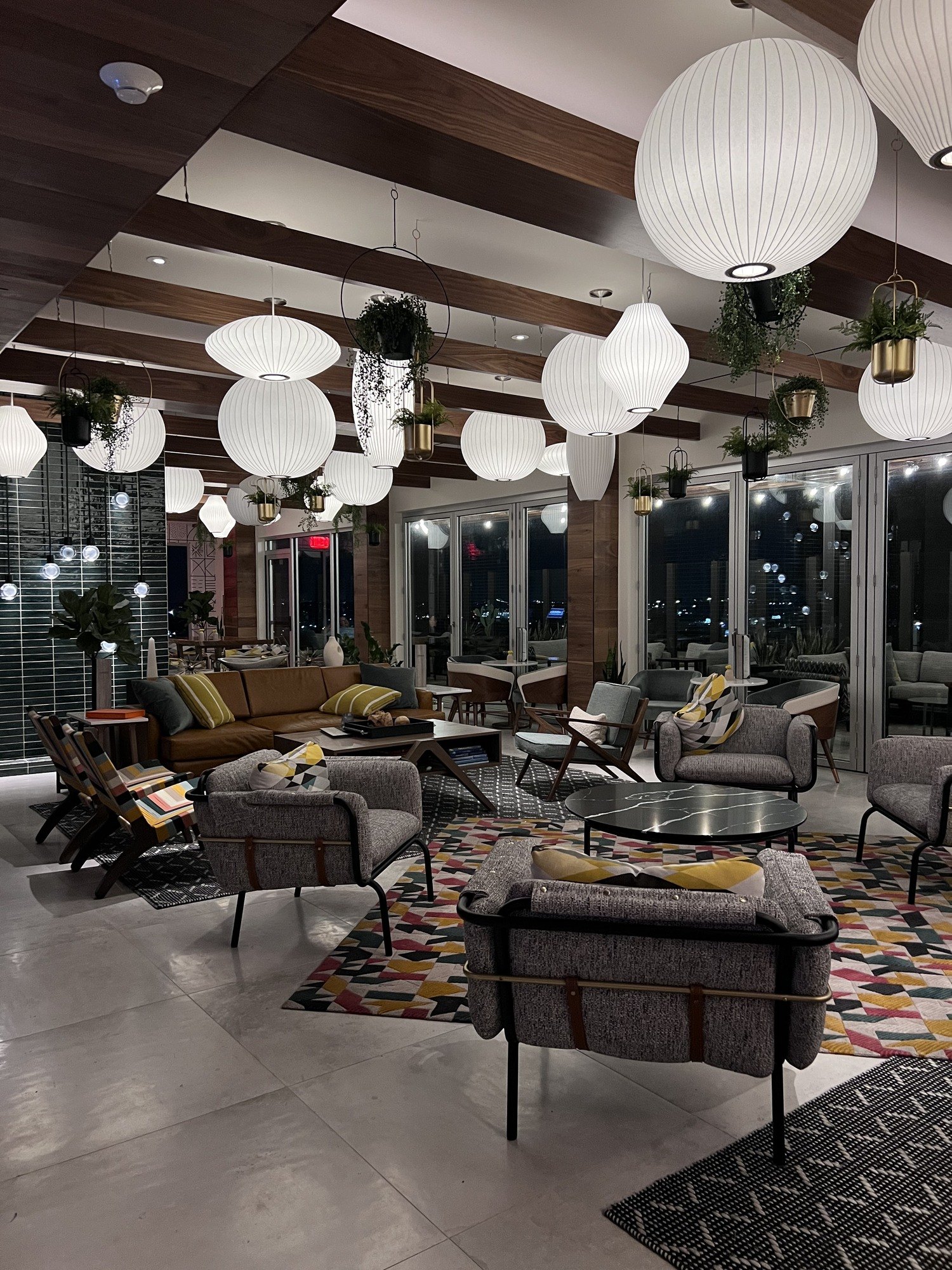 A lounge hung with white paper globes of different sizes under dark timber beams, with low armchairs on a patterned carpet and the city through the windows