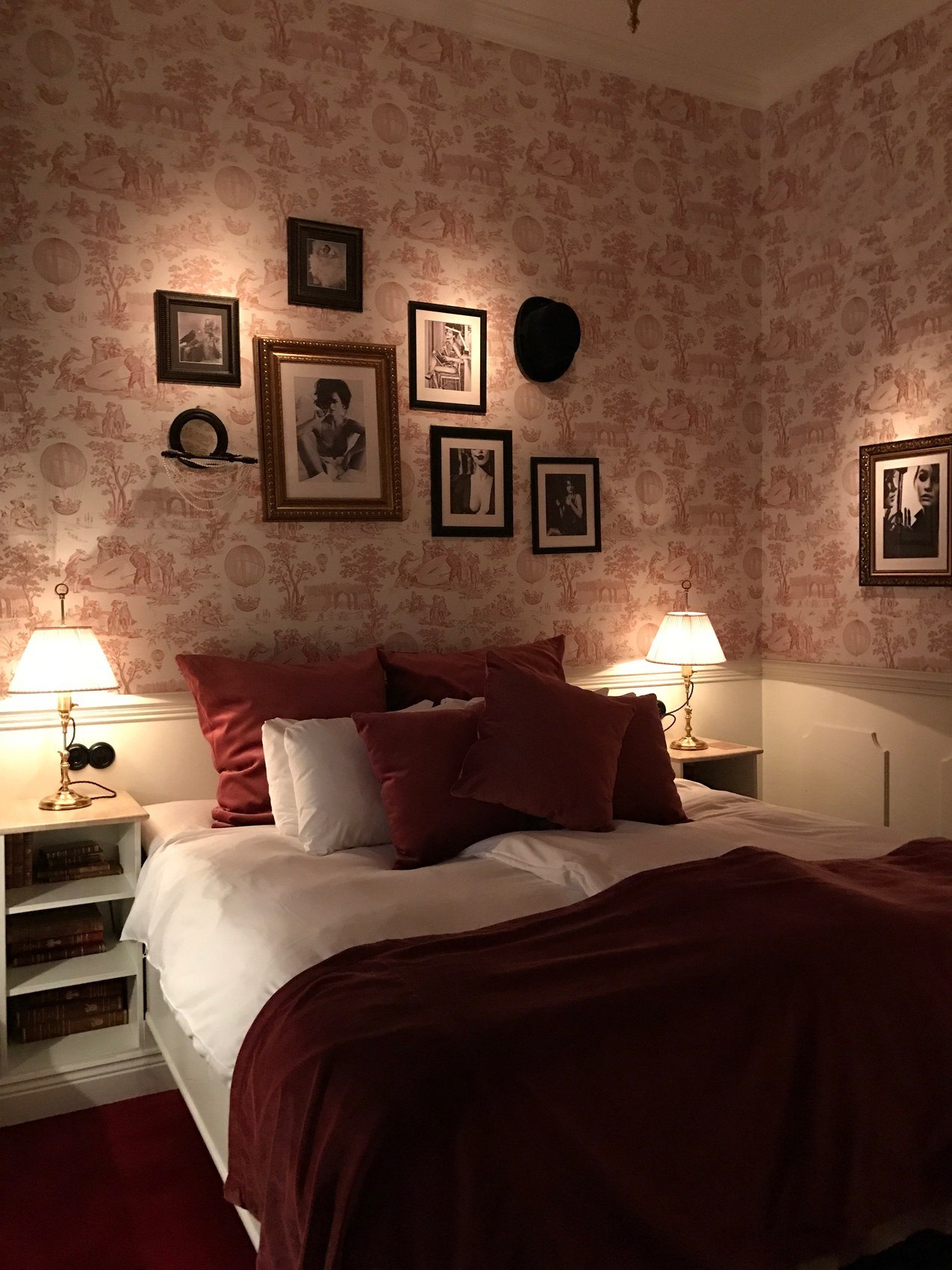 A bed in red and white under a wall of framed pictures on pink toile paper