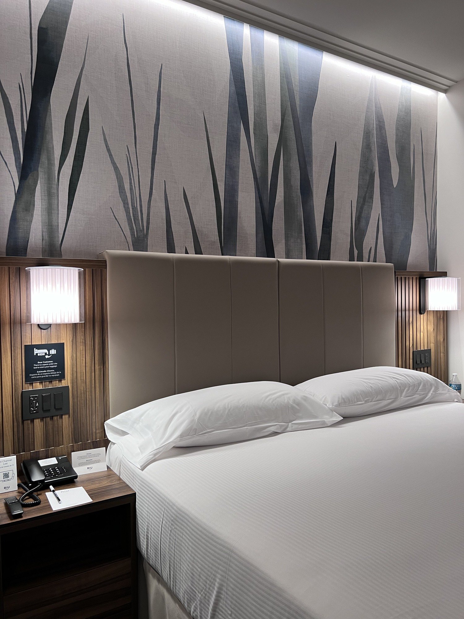 A bed with white linen against a tall headboard panel painted with pale grasses, a reading light on each side and a dark timber nightstand