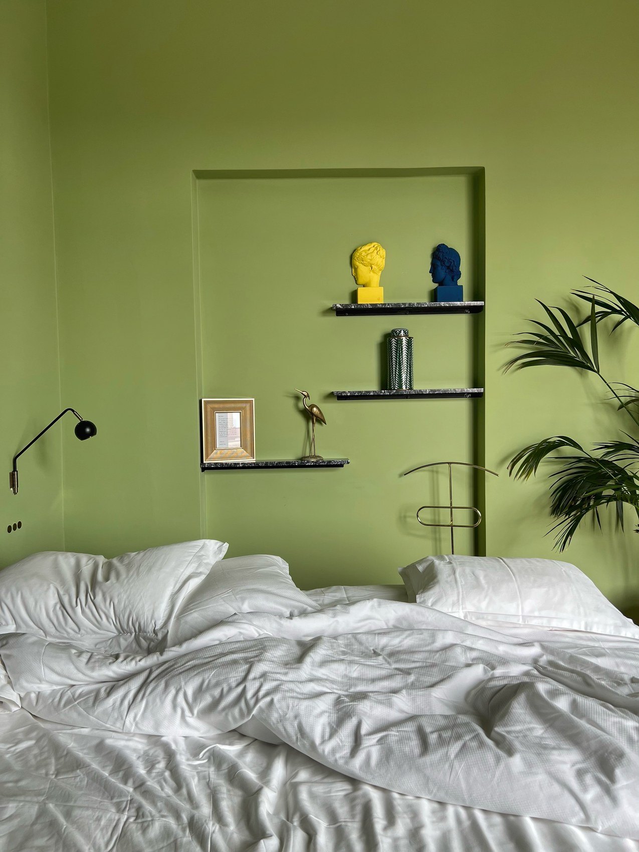 An unmade bed against an olive green wall with two small busts on a shelf