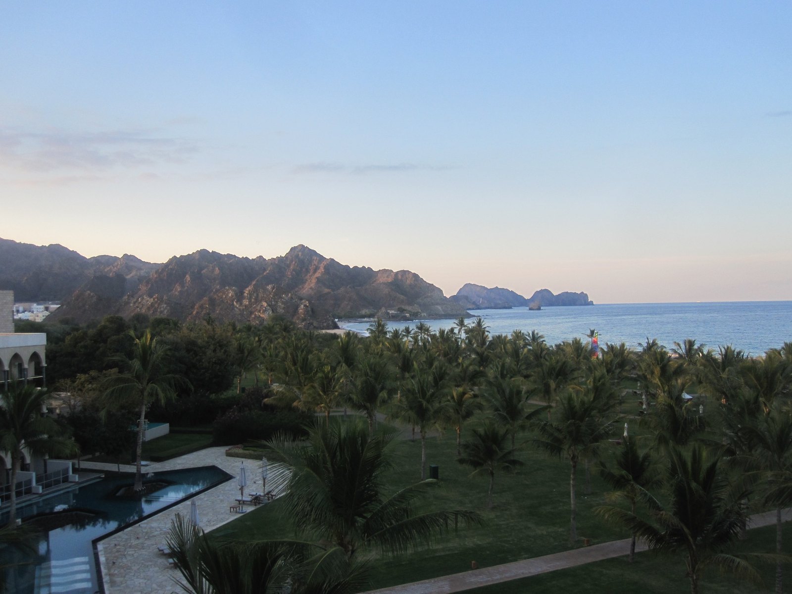 A palm garden and long dark pools below a ridge of bare Hajar mountains running down to the sea at dusk