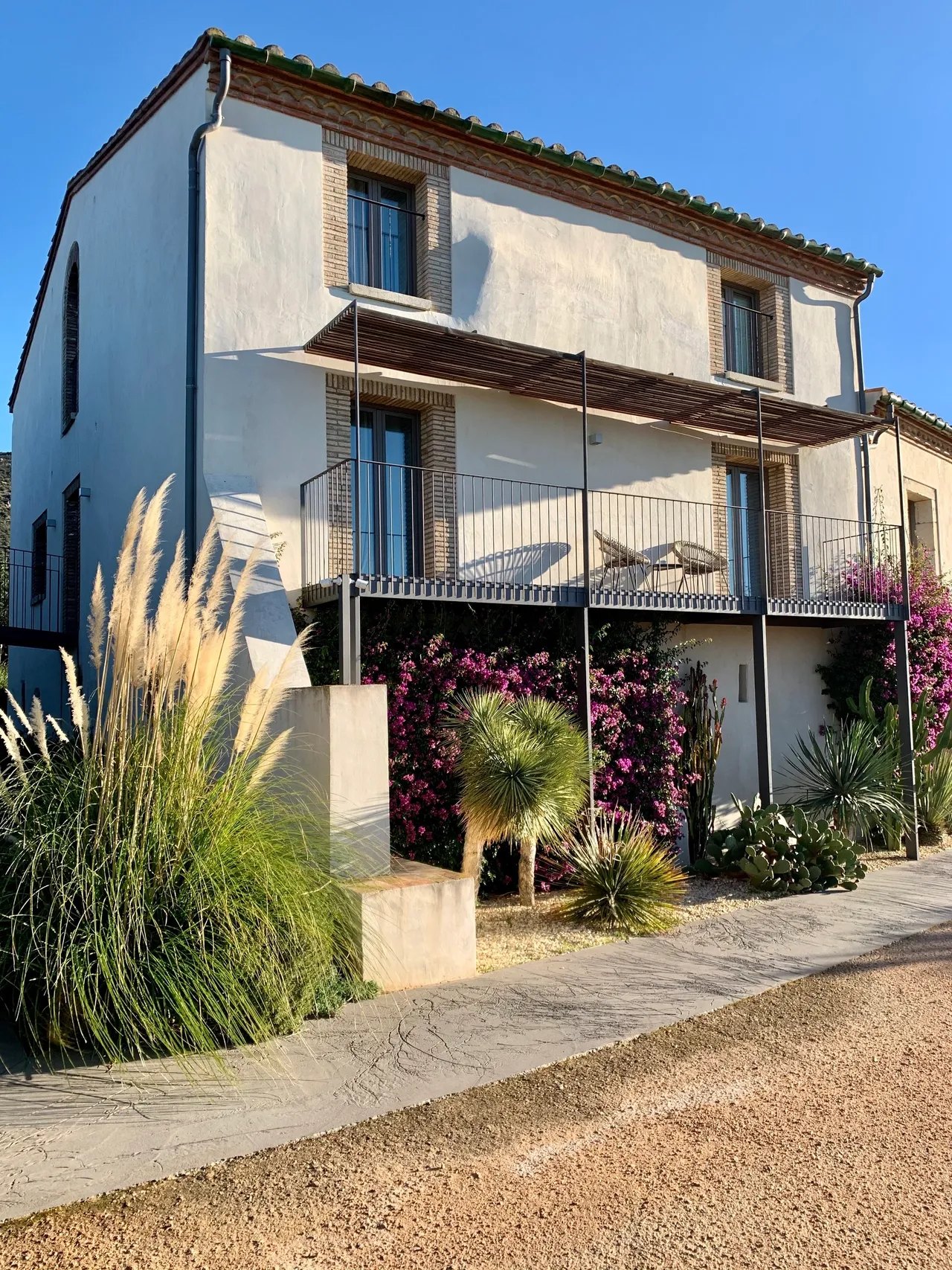 A rendered farmhouse with brick window surrounds and a green glazed tile roof, a slatted pergola shading the first floor terrace, with bougainvillea, pampas grass, yuccas and prickly pear in the gravel below