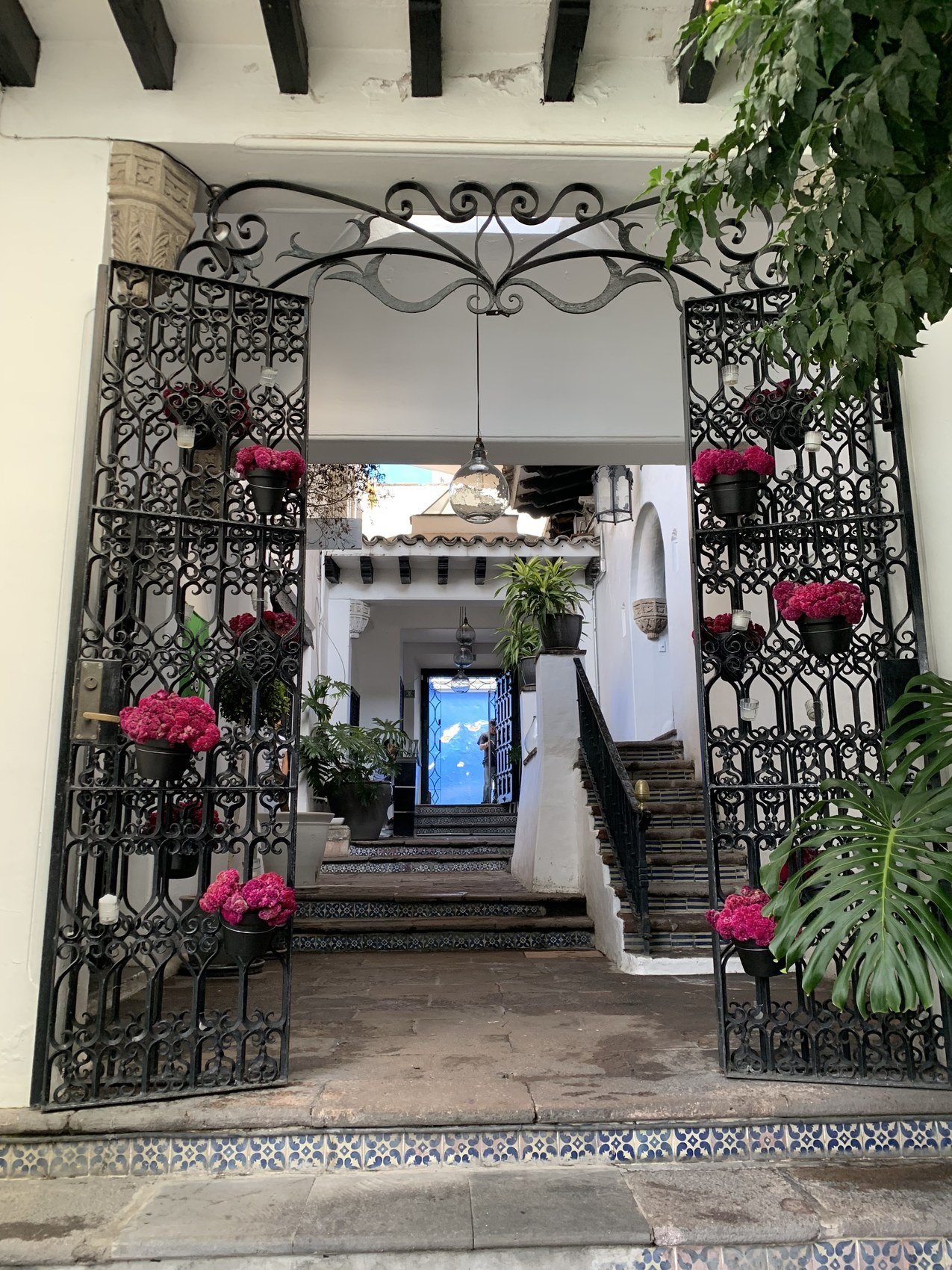 A wrought iron gate hung with pots of pink flowers, steps climbing into the house beyond