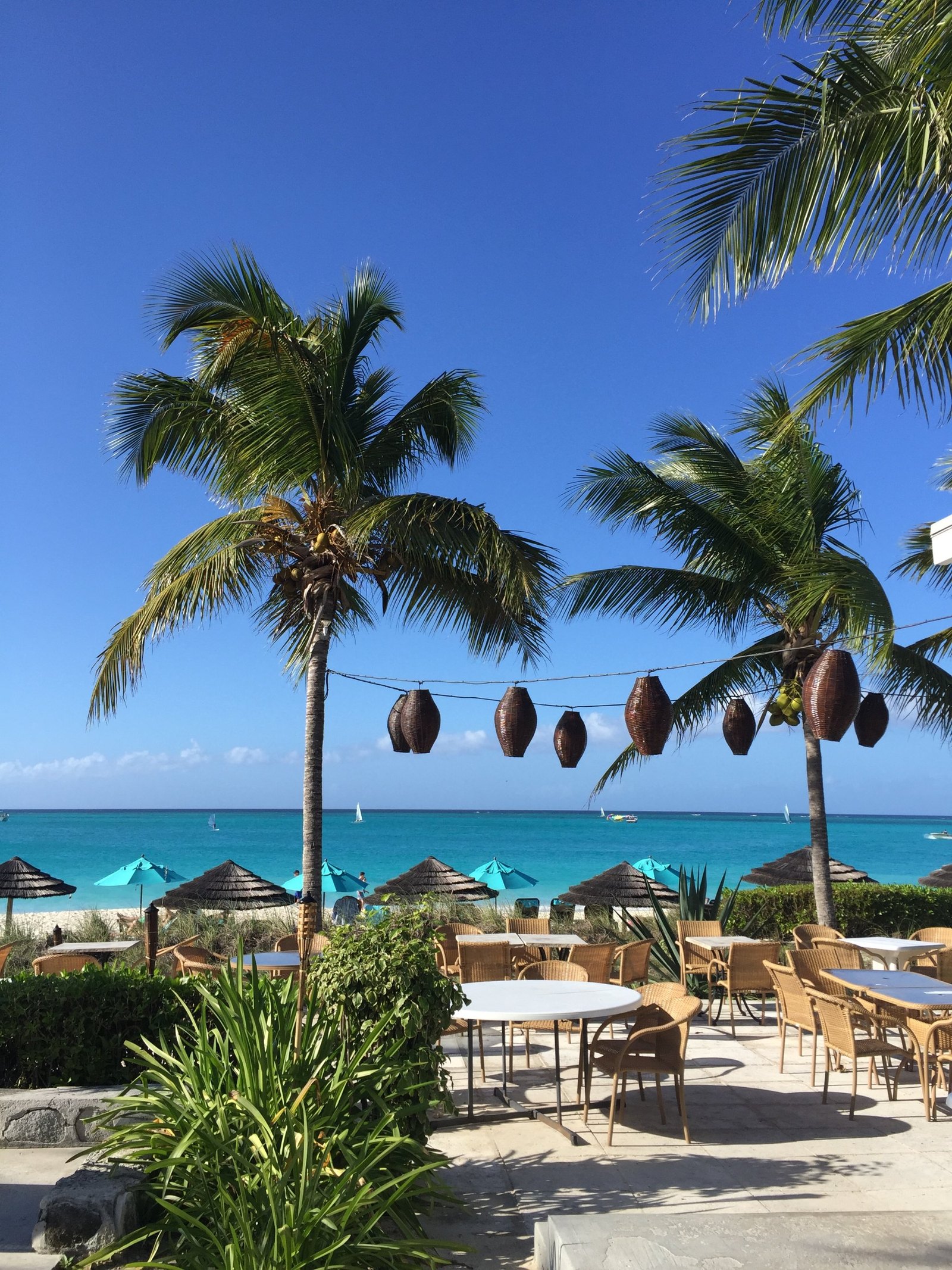 Woven lanterns strung between two palms over a terrace of wicker chairs, turquoise parasols on the sand beyond