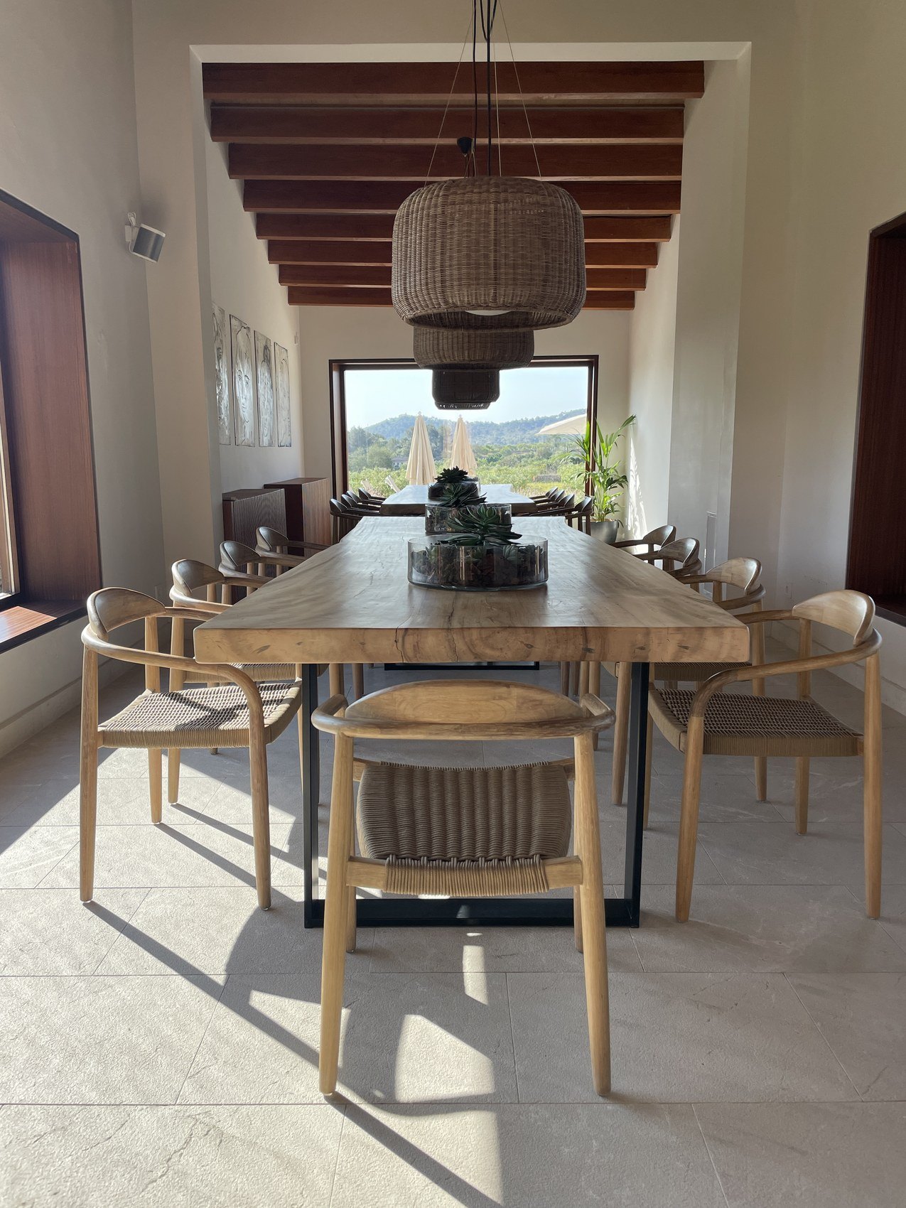 A long timber table and pale chairs under a beamed ceiling with the hills through the window