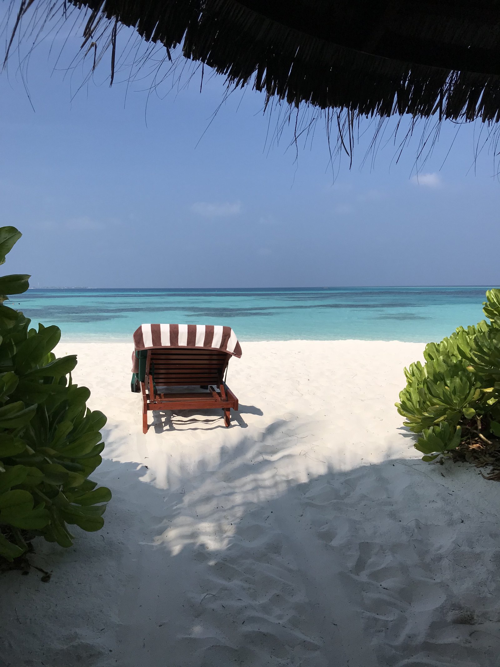 A single striped lounger on white sand framed by the thatch eave of a villa