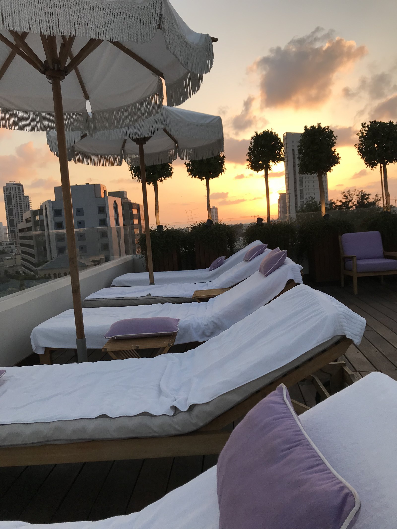 A row of timber daybeds with lilac cushions under fringed parasols on a roof at sunset