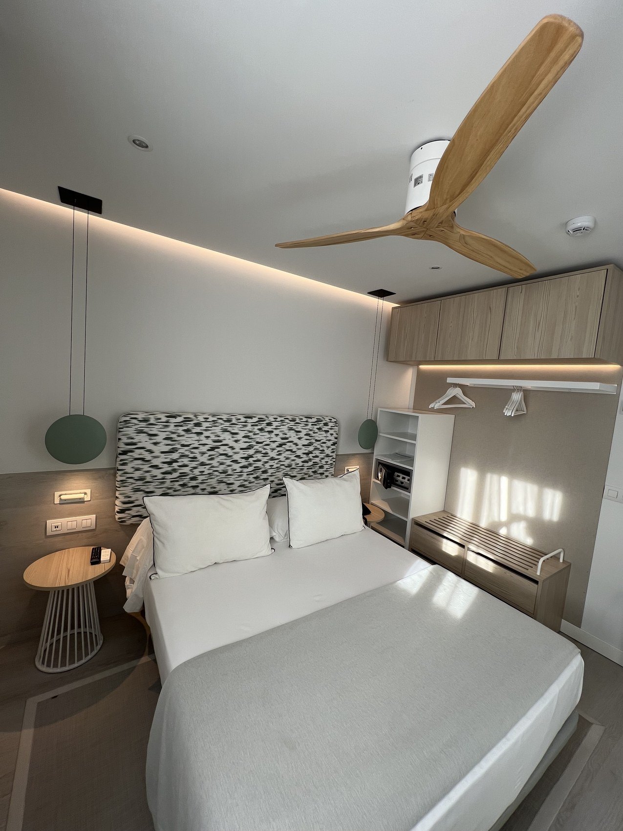 A pale room with a patterned headboard, a timber ceiling fan and built-in shelving