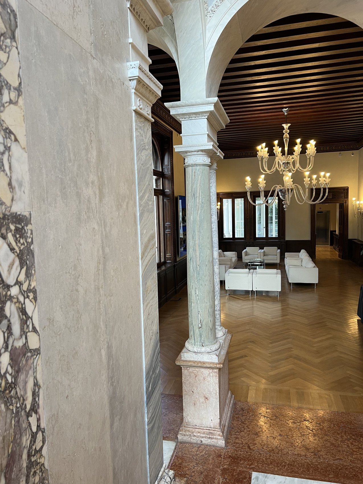 A marble column under a dark coffered ceiling, with a chandelier and white sofas beyond