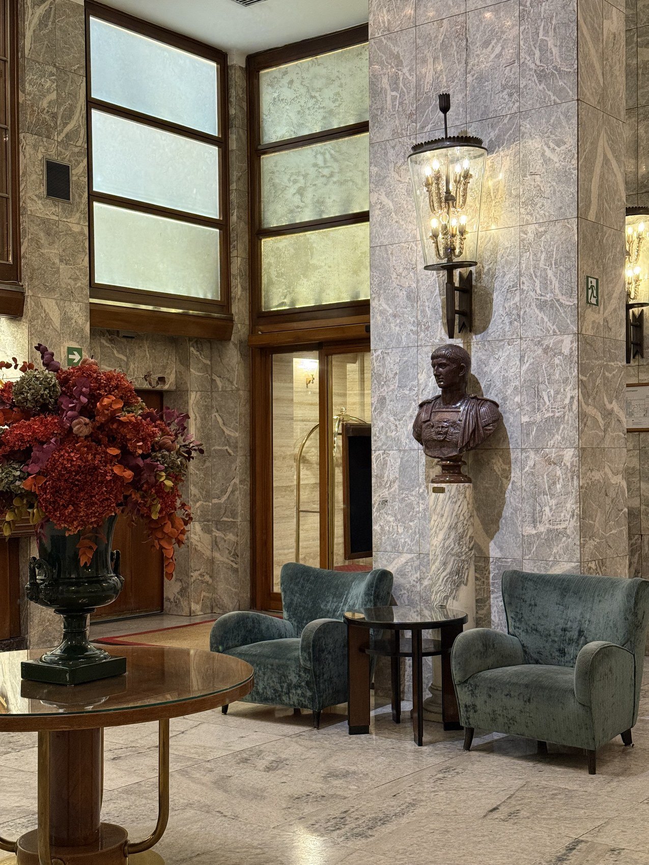 A marble lobby with a bronze bust on a column, green armchairs and an urn of autumn flowers