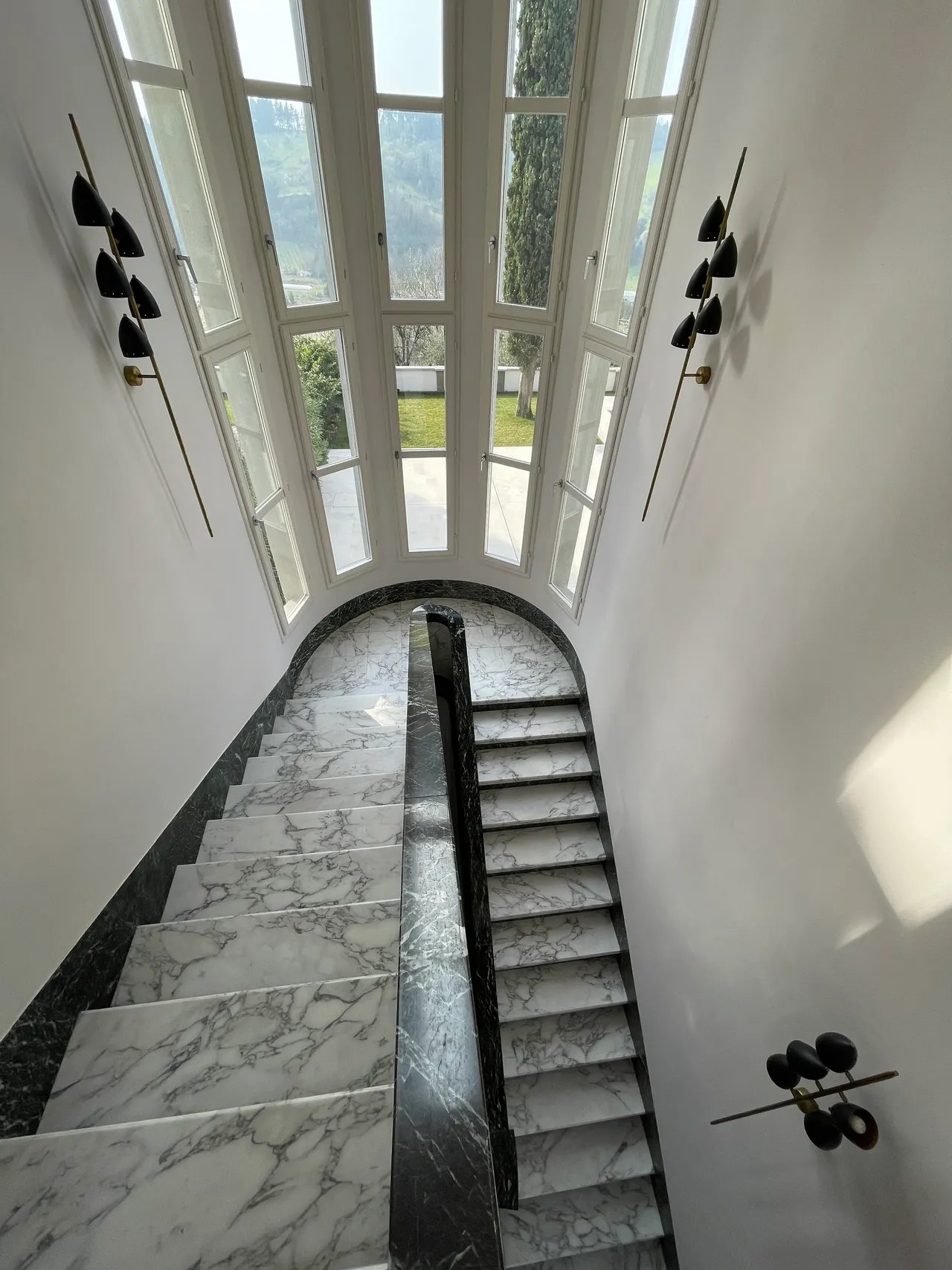A staircase of white and dark green marble seen from above, running down to a tall many-paned arched window with a cypress and green hills beyond, black and brass sconces on the walls