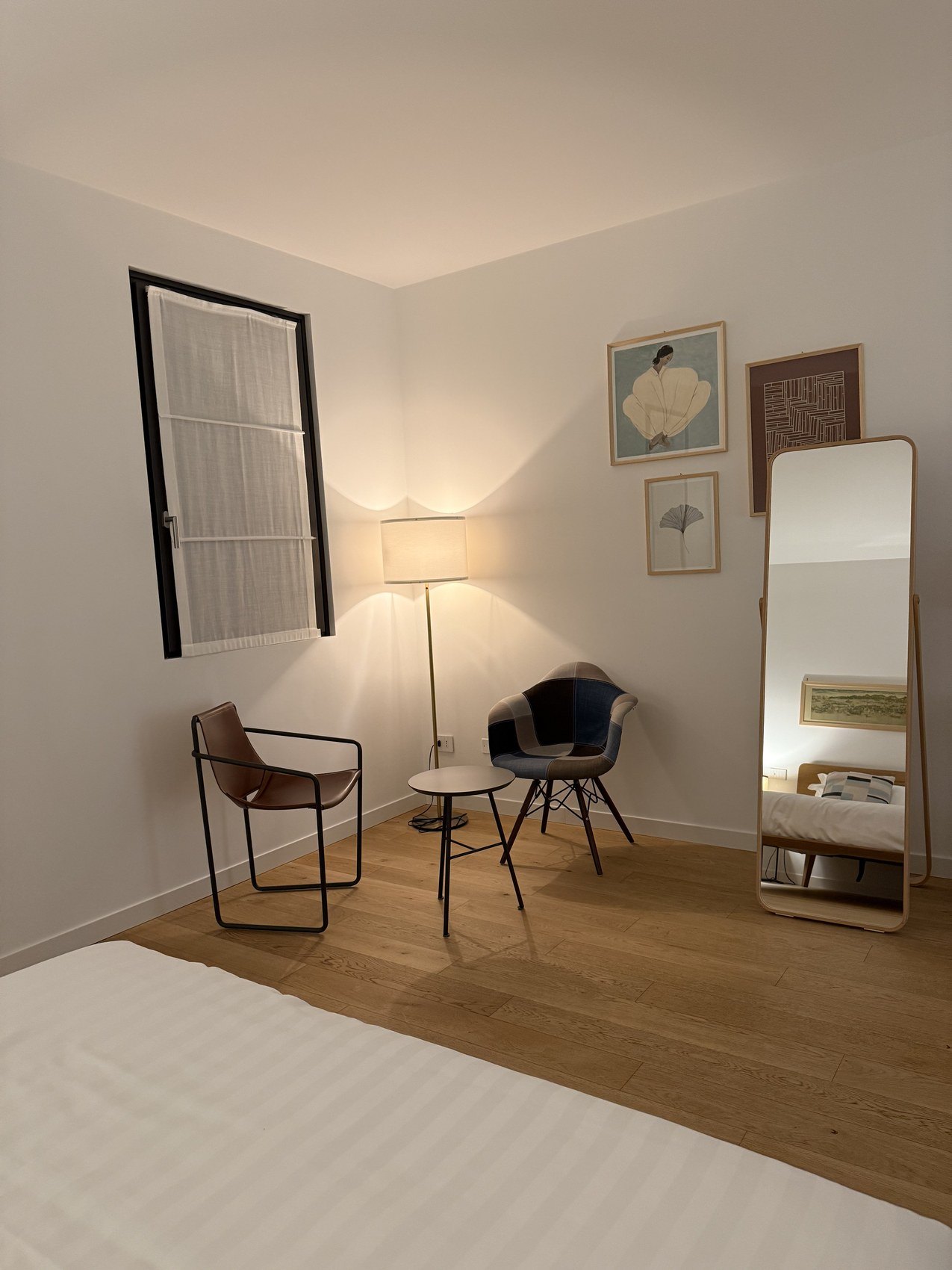 A full length mirror beside two moulded chairs and a floor lamp on a pale timber floor