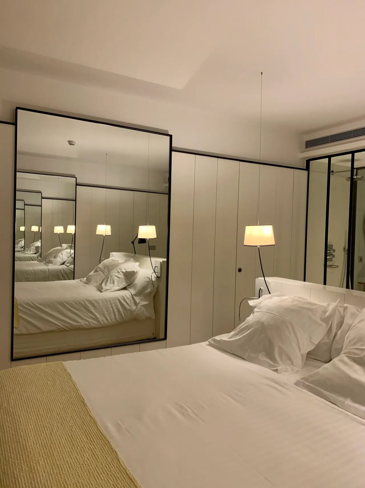 A bedroom with a tall black framed mirror doubling the bed, a pendant lamp hanging on a long flex, pale panelled wardrobes and a glazed partition through to the shower