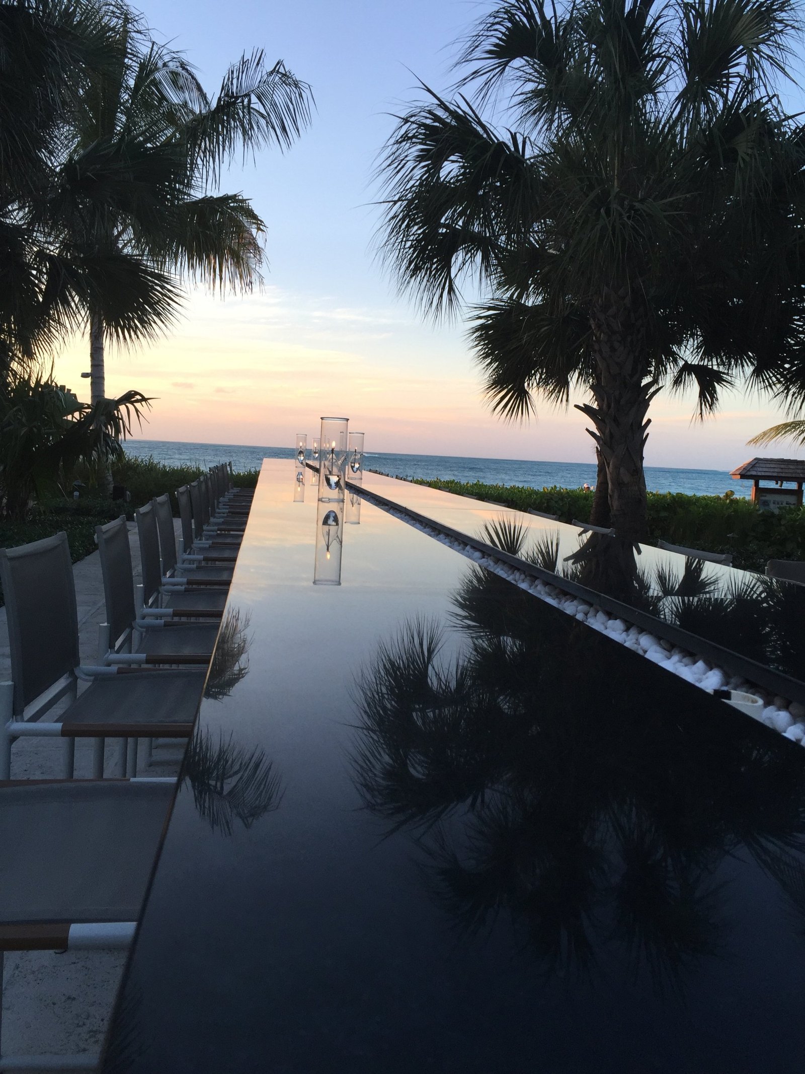 A long dark reflecting table set with glass hurricane lamps, palms and the sunset doubled in its surface