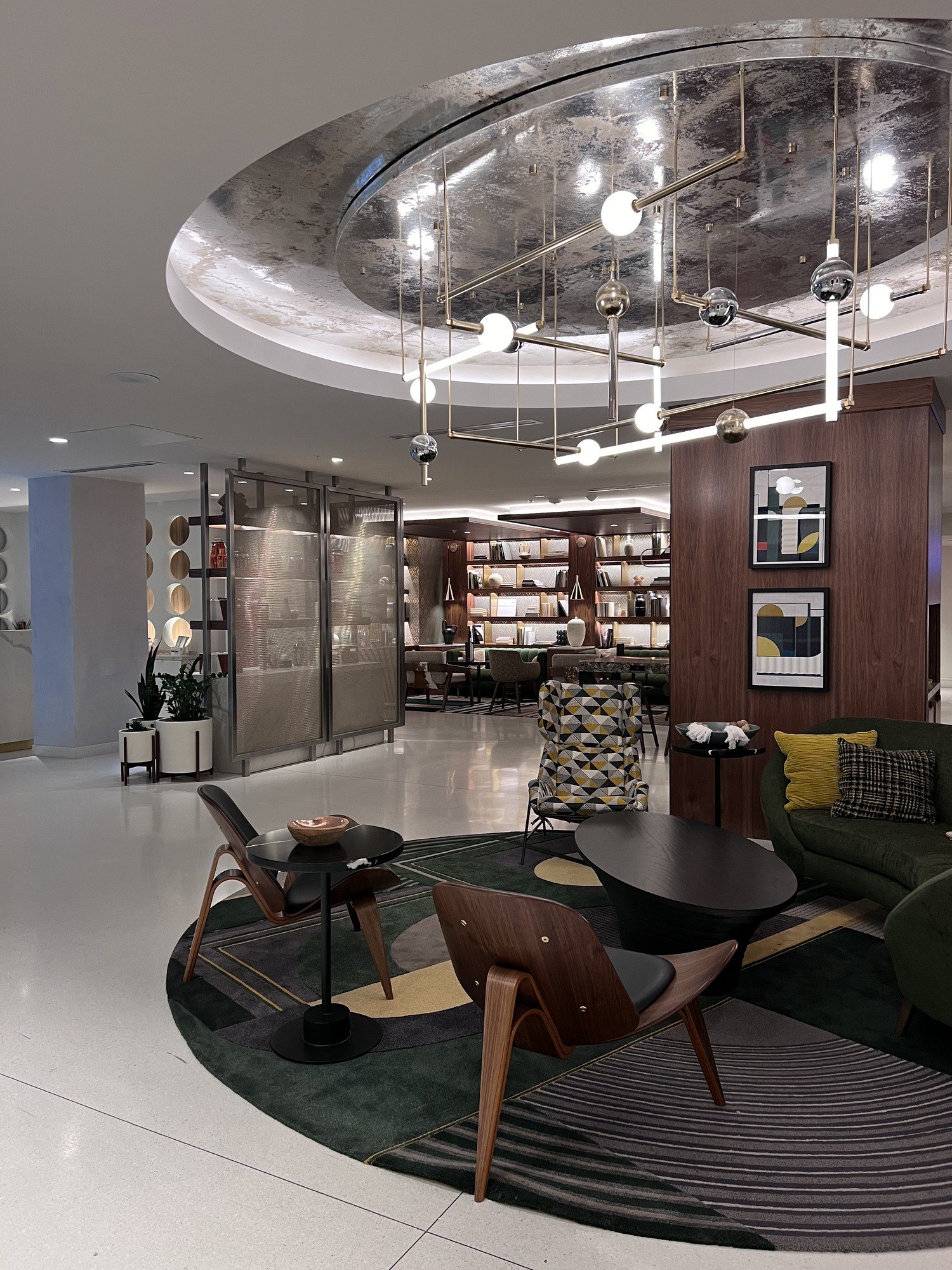 A hotel lobby under a large mirrored ceiling disc hung with scattered bulbs, with mid-century timber chairs on a dark green rug and shelves of books along the back wall