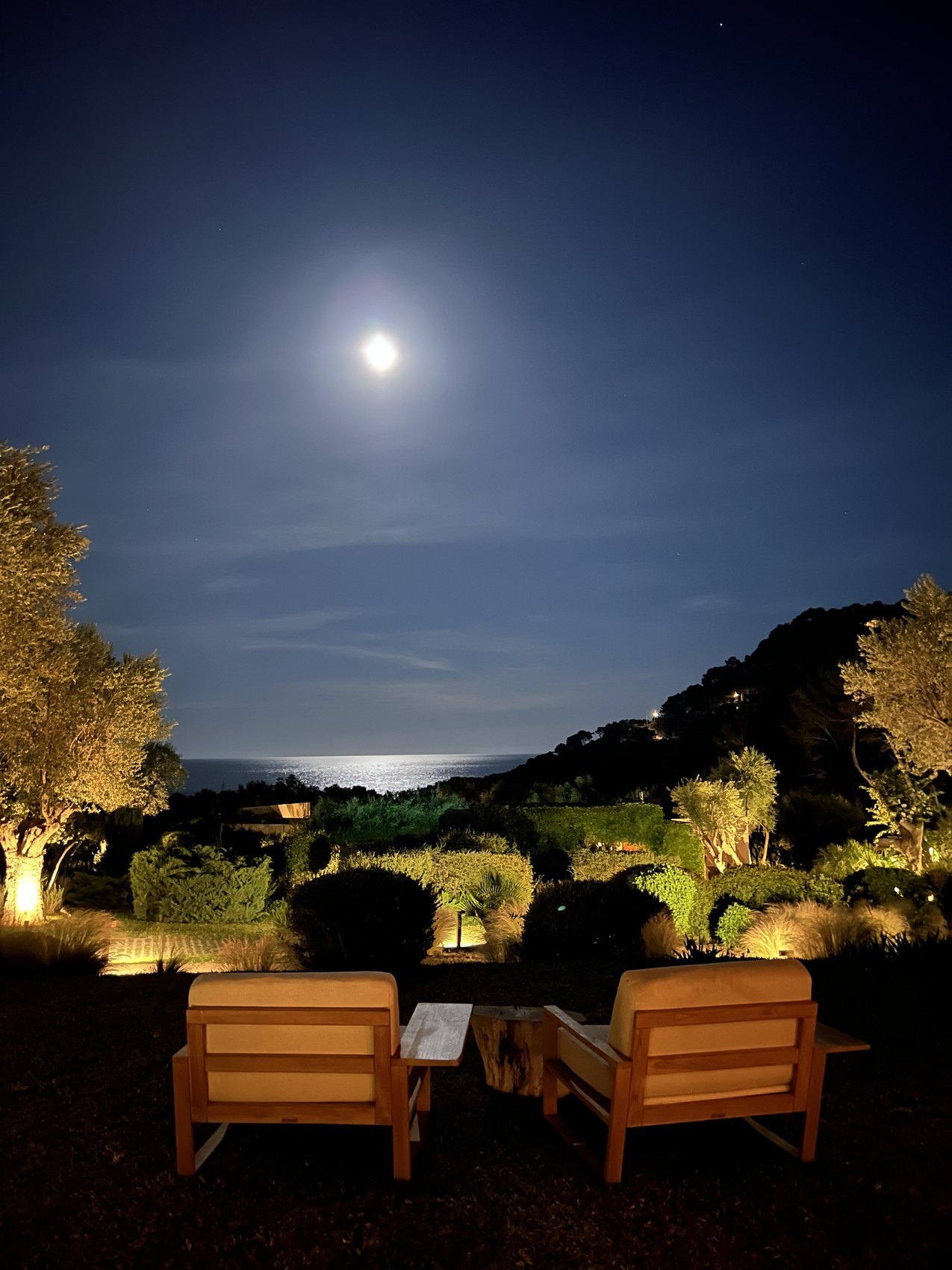 Two timber armchairs lit on a terrace at night with the full moon over the sea beyond the garden