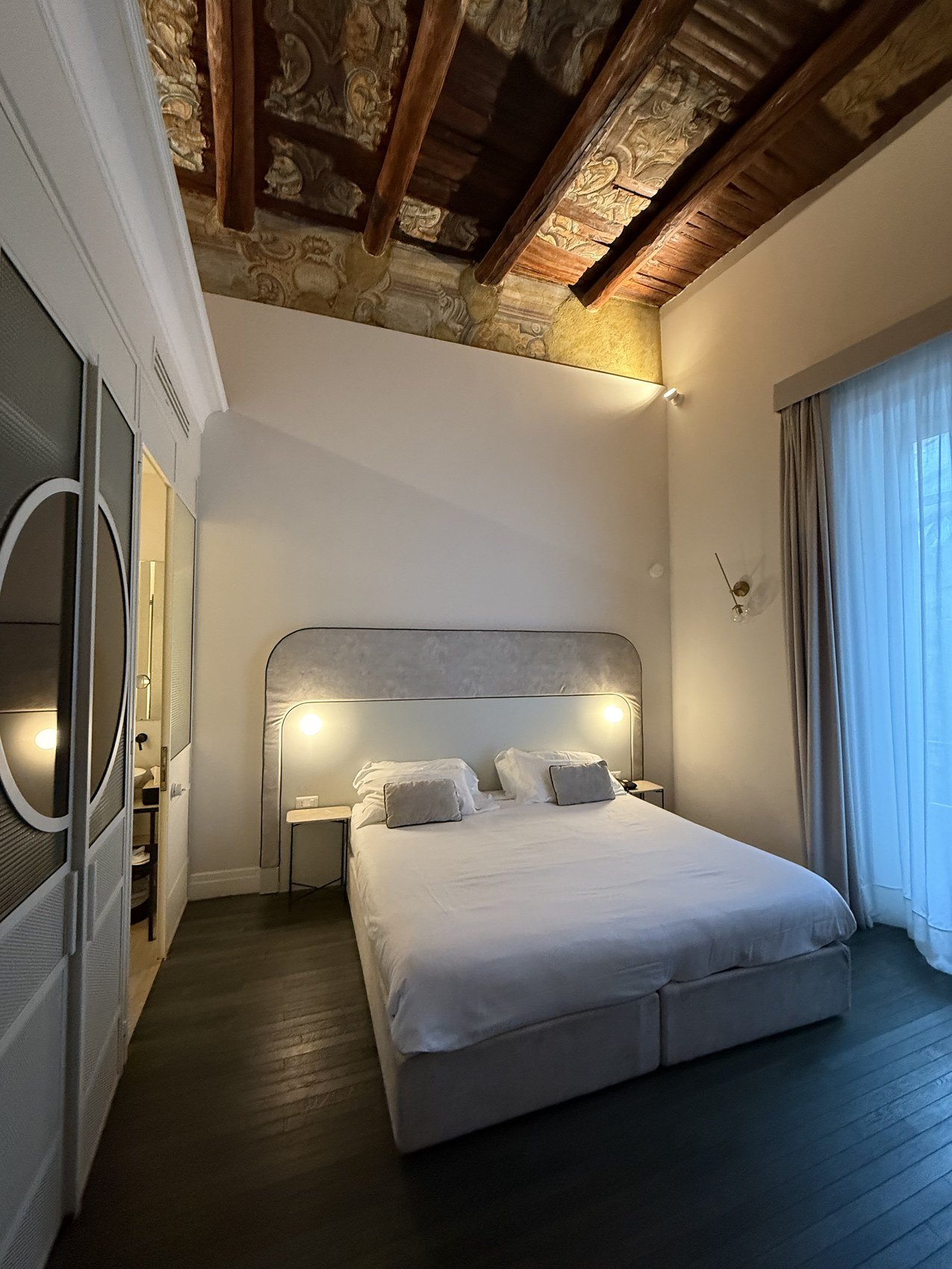 A bed in a white room under an old beamed ceiling with a rounded glass partition beside it