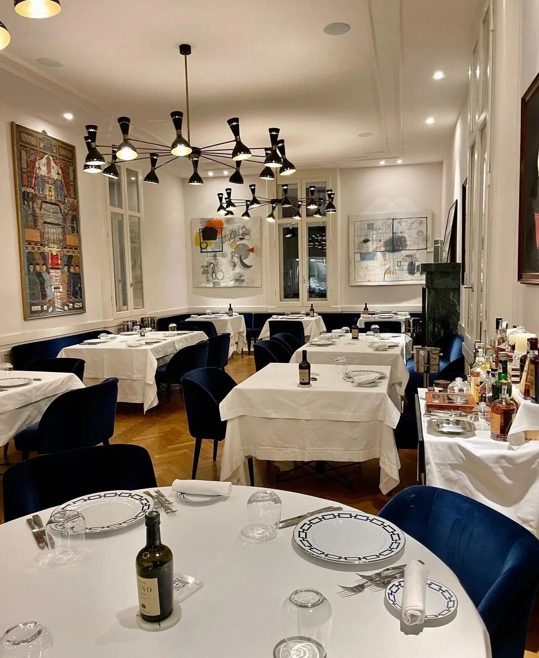 A dining room with white clothed tables and navy velvet chairs under two black many-armed chandeliers, a parquet floor, tall shuttered windows and framed artworks on the walls