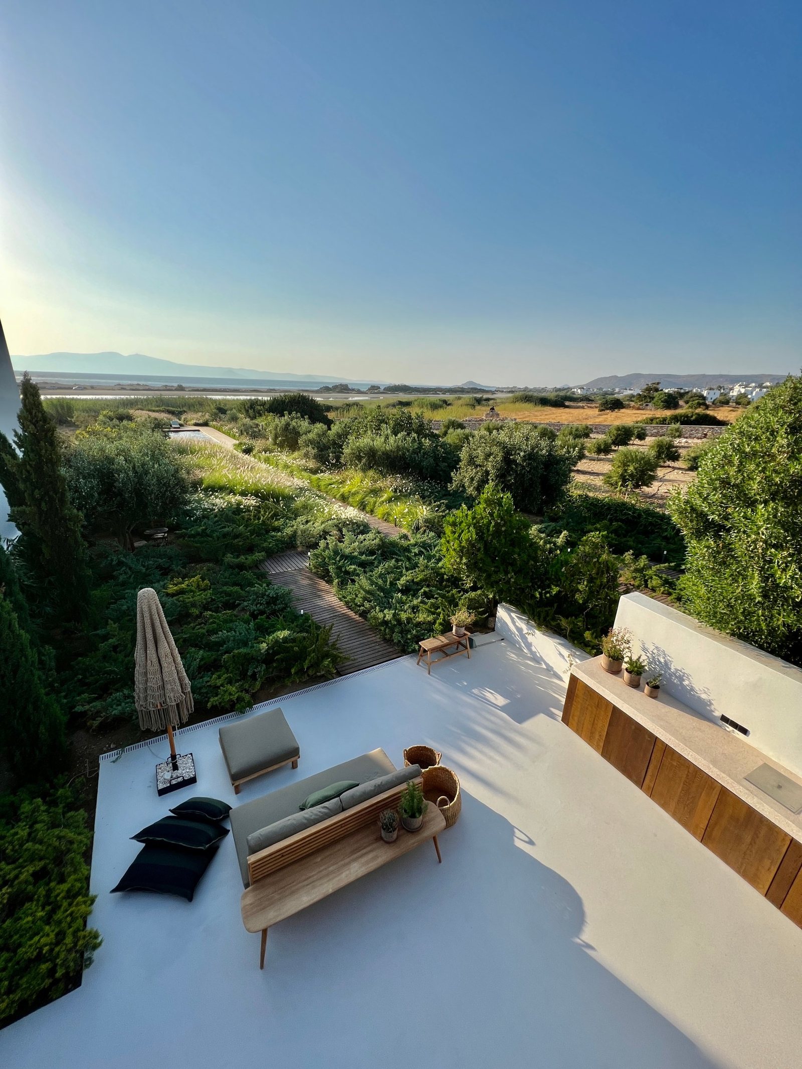 A white terrace with a parasol and an outdoor kitchen, the garden and the sea beyond