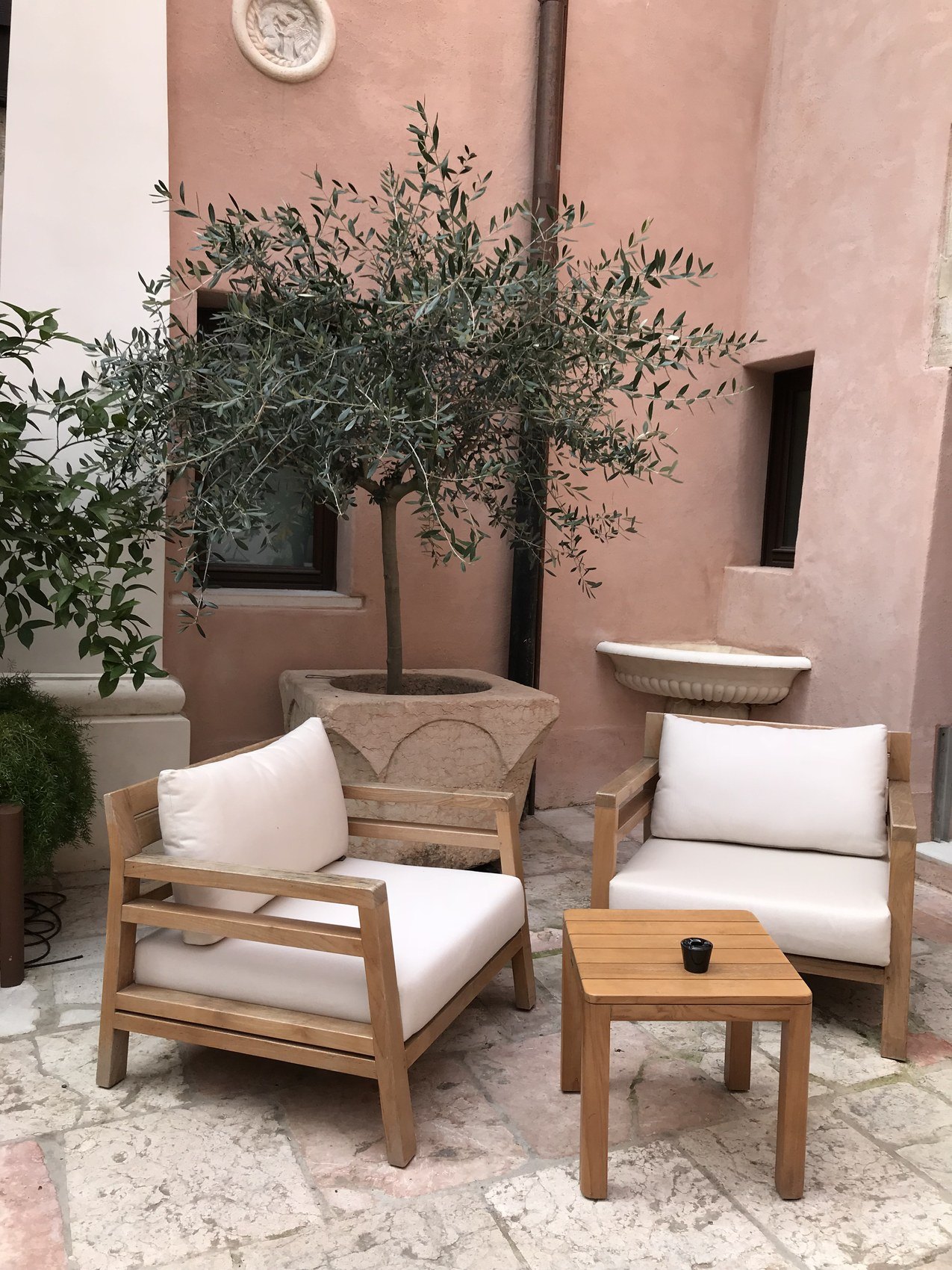 An olive tree in a stone trough in a pink walled courtyard with pale timber armchairs