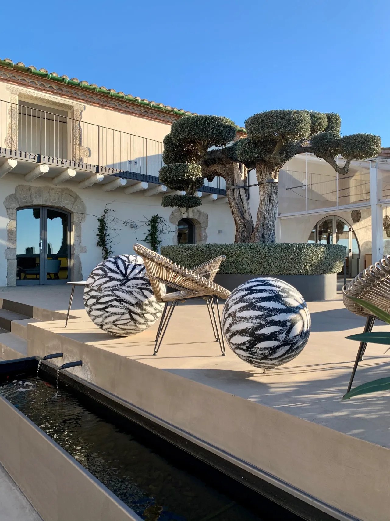 A courtyard with two clipped olive trees above a long hedge, large spheres painted in black and white brushstrokes and rattan chairs on pale paving, a narrow water channel in the foreground