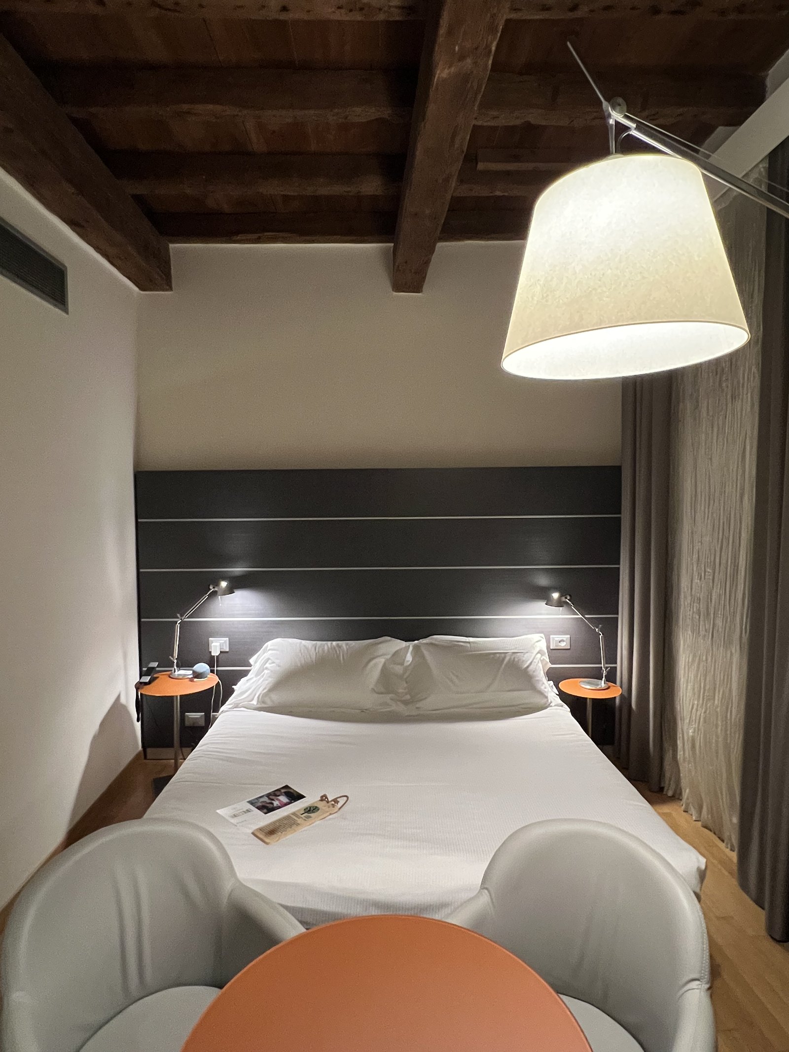 A bed under a dark beamed ceiling with a grey padded headboard panel and a lamp with an orange shade on each side, two white moulded chairs and an orange table in the foreground