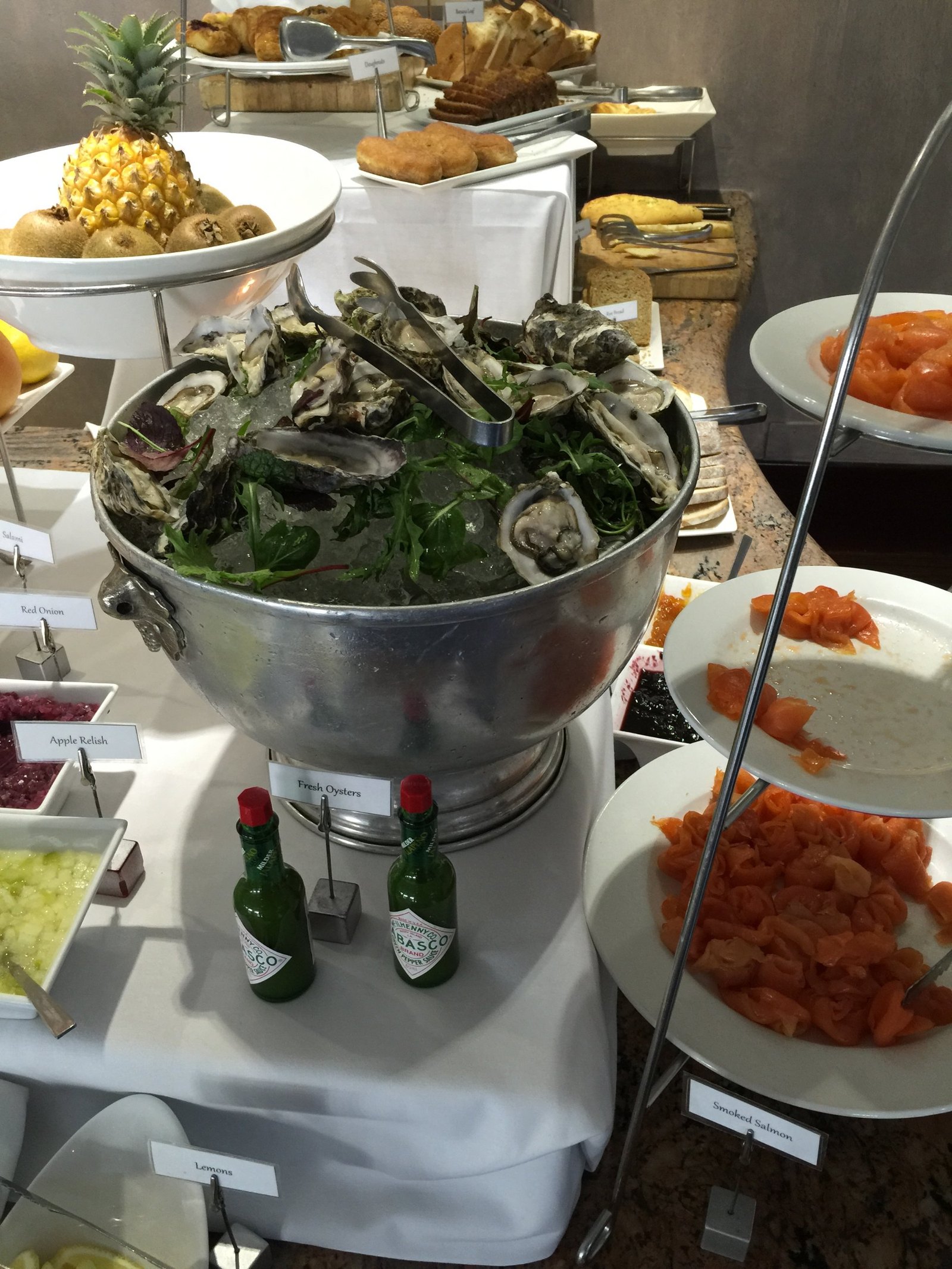 A breakfast buffet with a steel bowl of fresh oysters on ice, smoked salmon on two tiers beside it and a whole pineapple on a stand behind