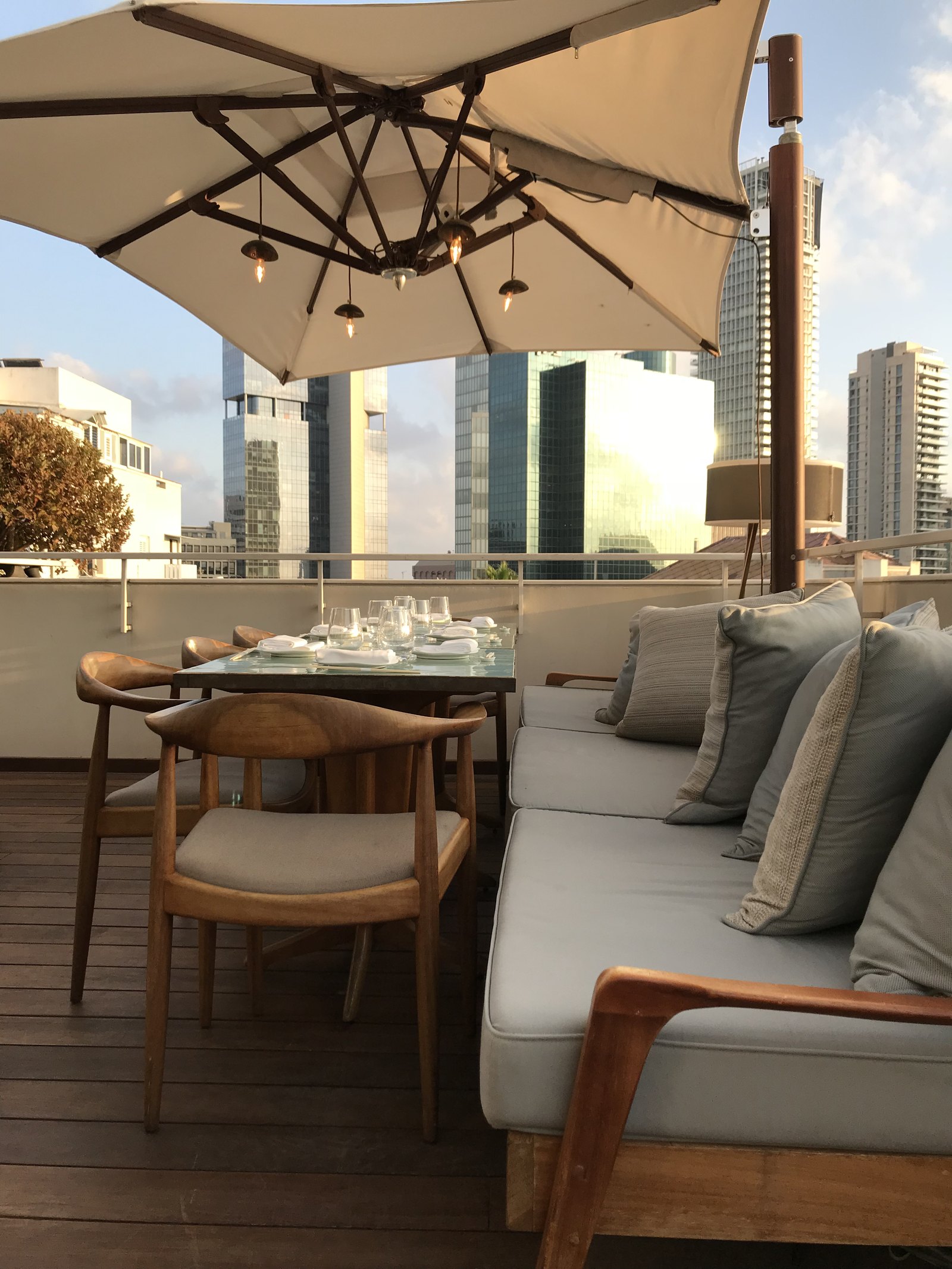 A laid table and a low sofa under a canvas parasol on a terrace, the glass towers of the city behind