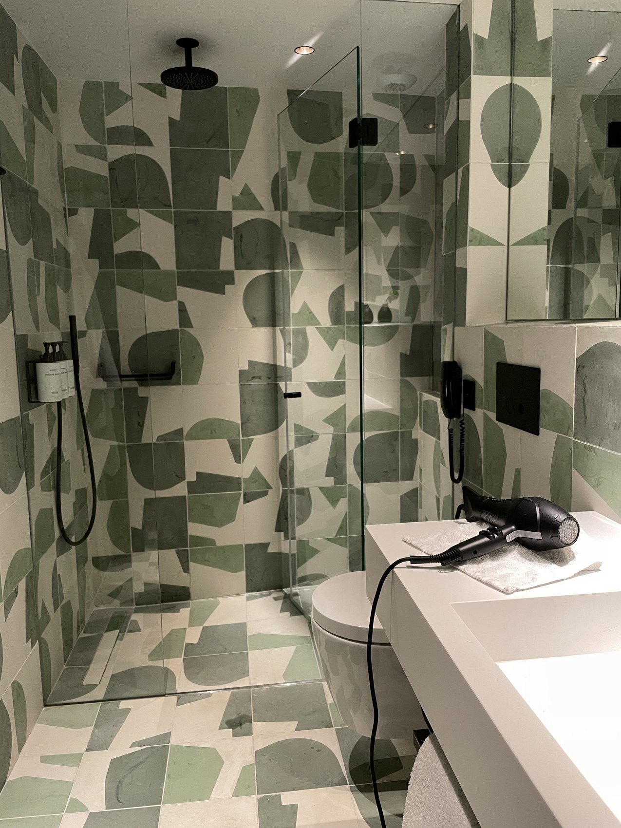 A walk-in shower tiled in a green and white abstract pattern behind glass