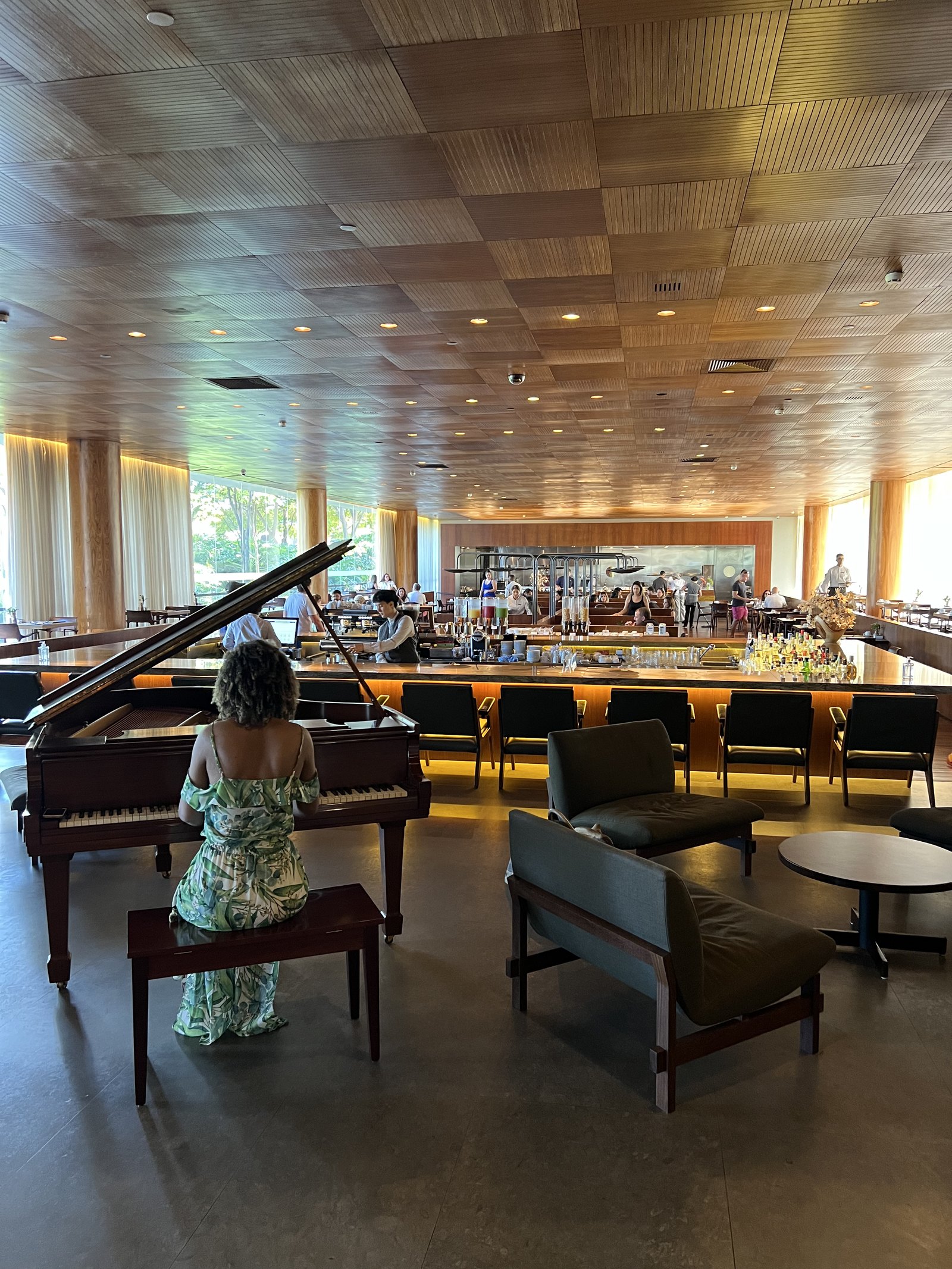 A woman in a long dress playing a grand piano in a tall lobby bar, under a timber slatted ceiling and a cloud-shaped light, with the bar running along the back wall