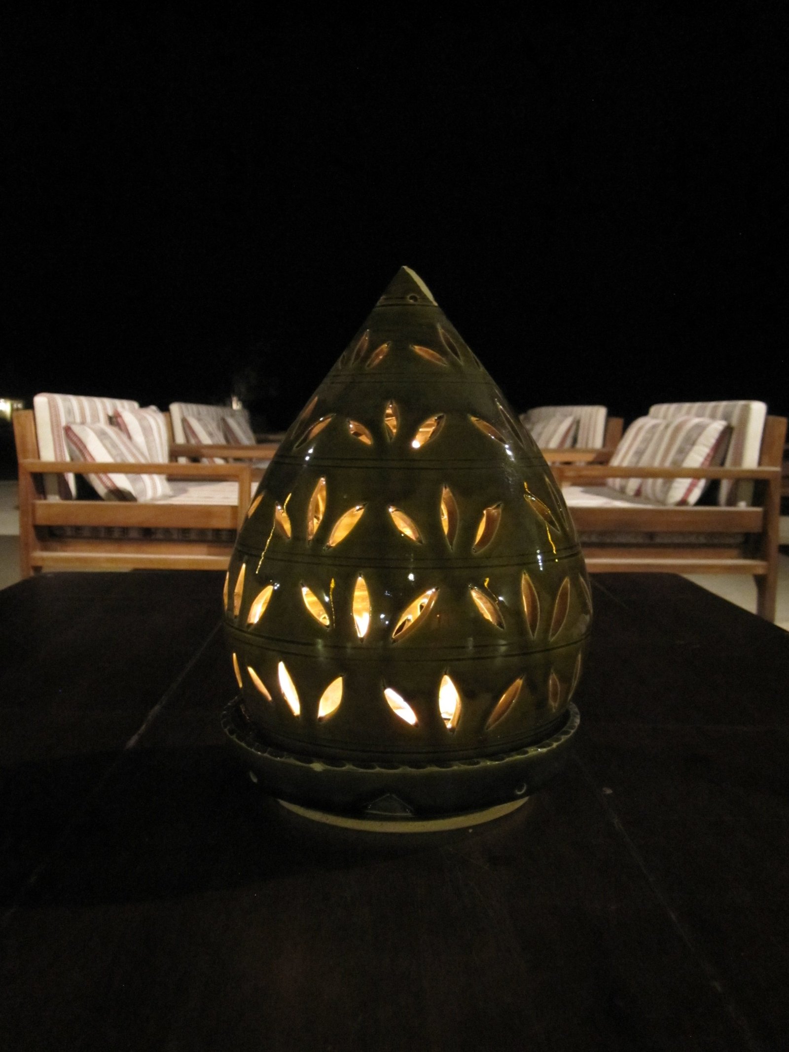 A green glazed lamp pierced with leaf shapes and lit from inside, on a dark table in front of striped cushioned chairs at night