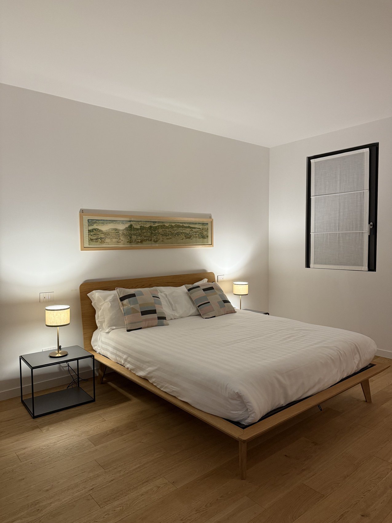 A bed with striped cushions between two small lamps under a framed landscape