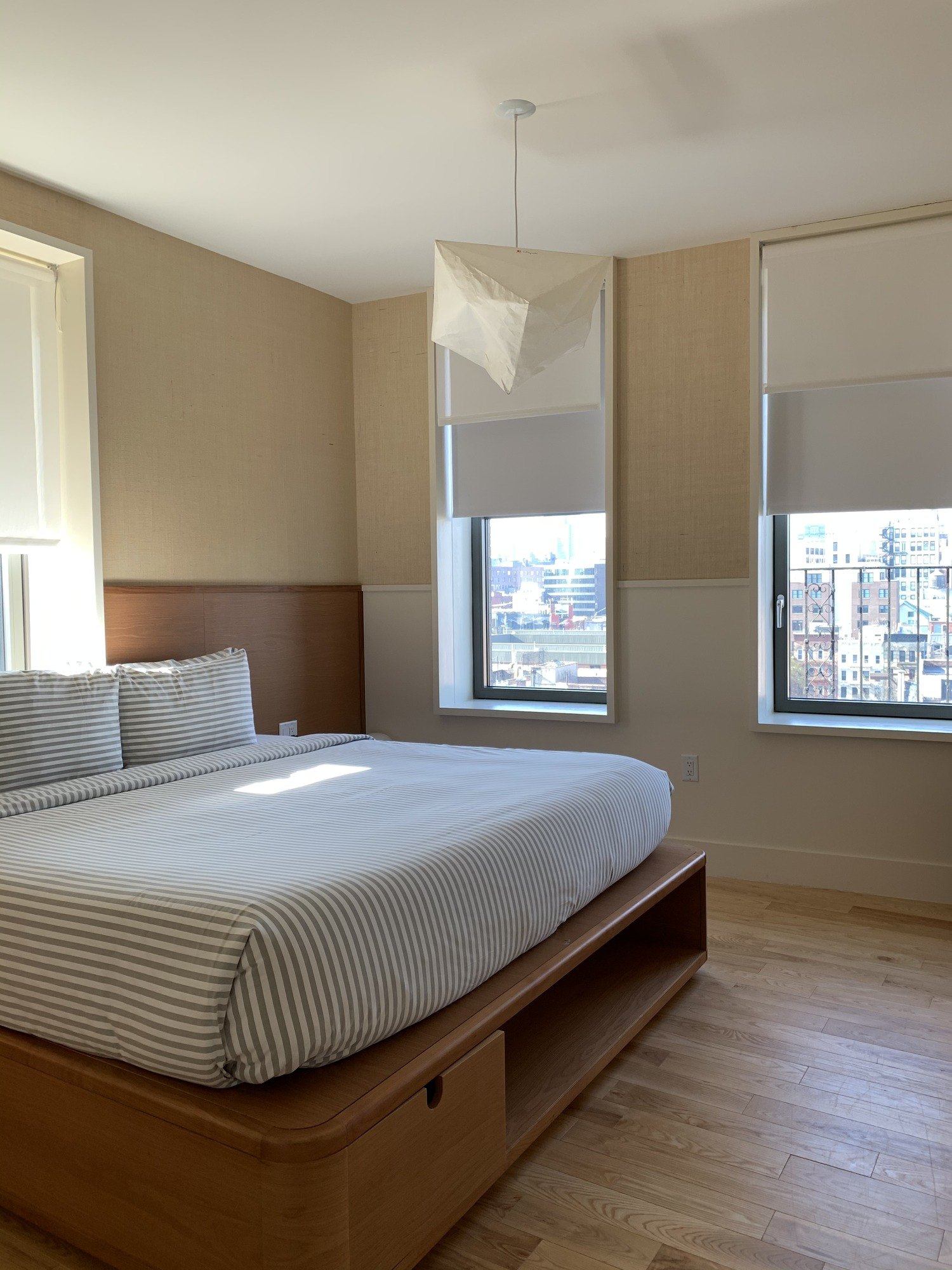 A corner bedroom with a timber platform bed under a striped cover, a paper pendant above and two tall windows onto the city