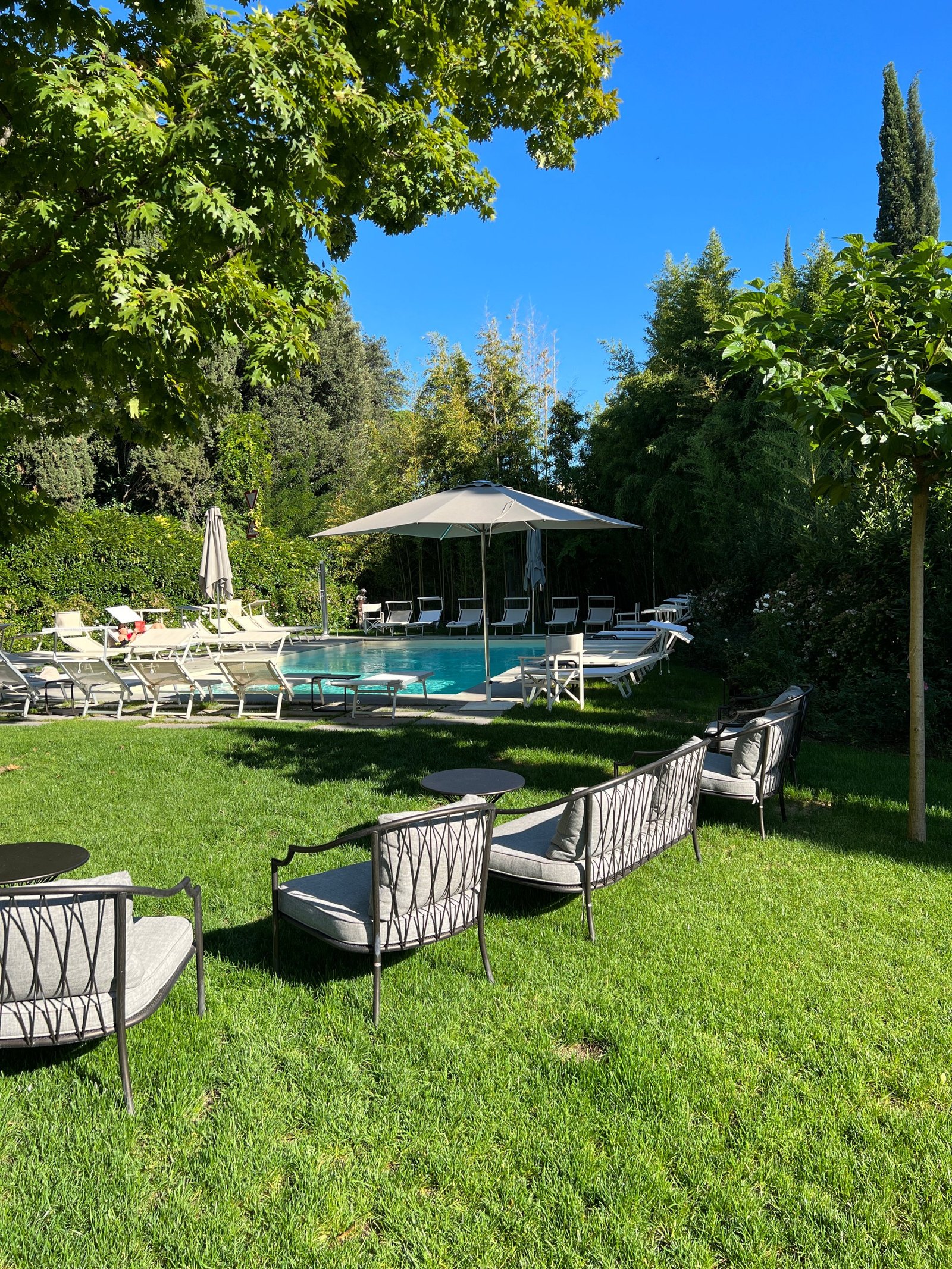 A pool with white loungers and a grey parasol at the far end of a lawn, with woven iron garden sofas in the foreground and cypresses and a plane tree around it