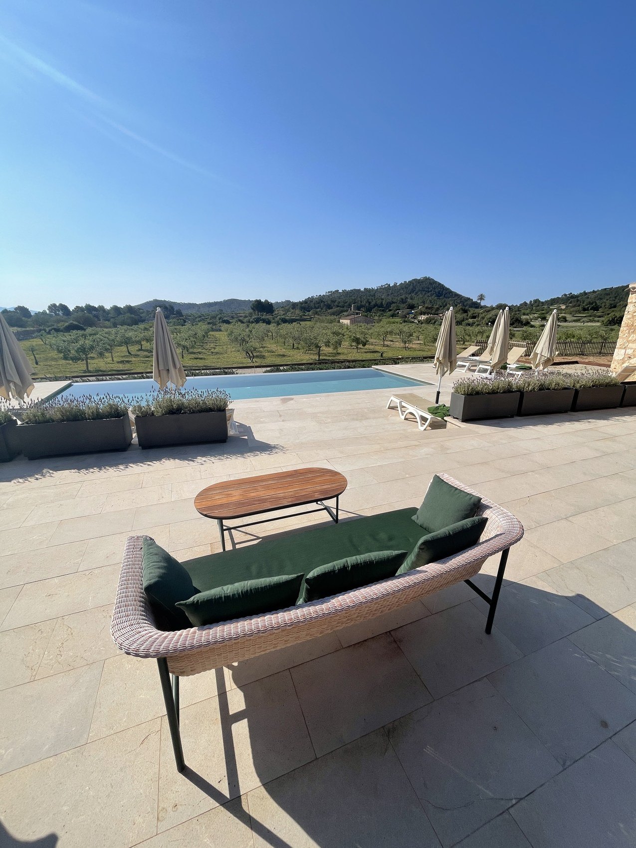 A stone terrace with a green cushioned daybed beside a pool, fields and hills beyond