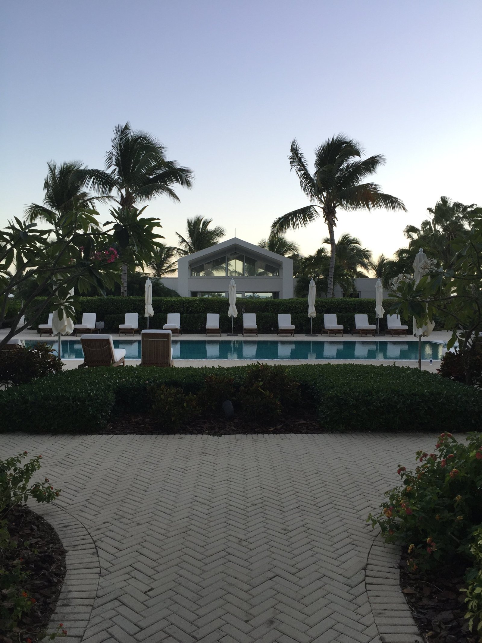 A long pool at dusk with a row of white loungers and a gabled glass fronted pavilion behind it